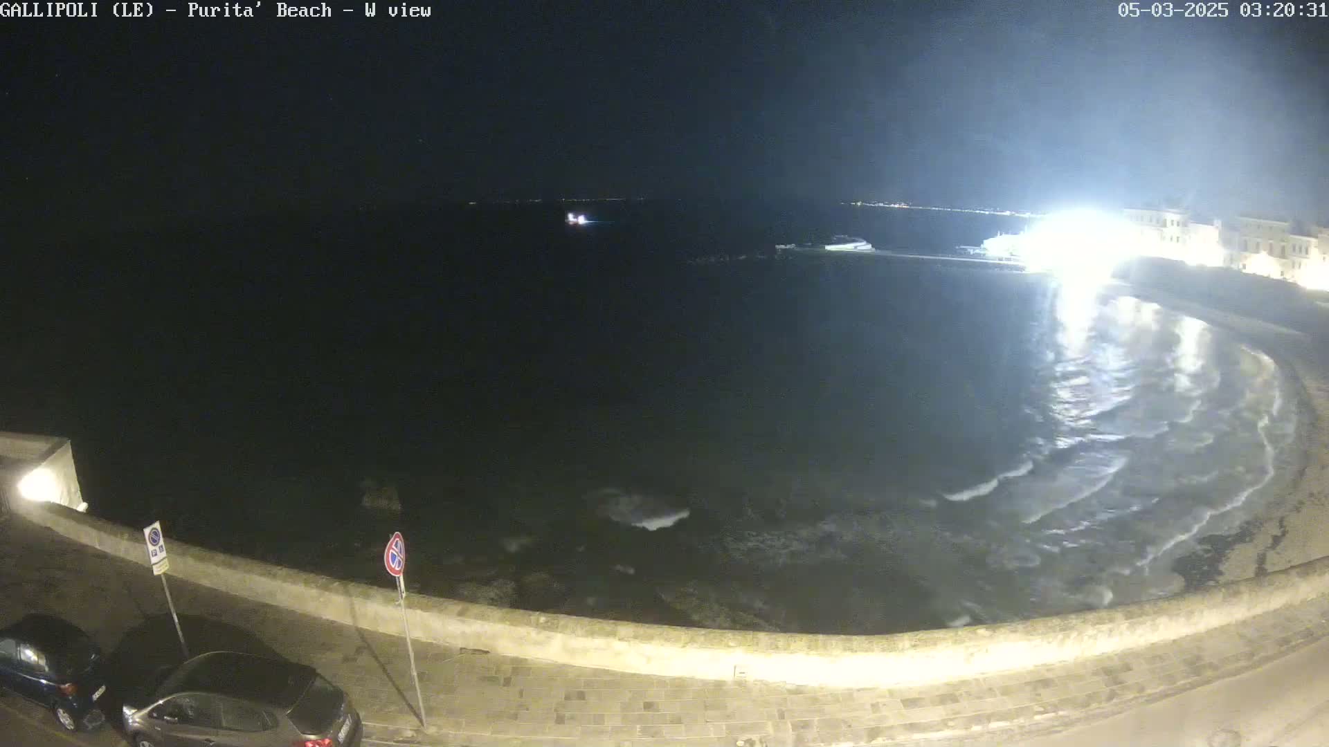 A nighttime, high-angle view of a dark, wavy sea lapping against a stone wall, with two cars parked along the wall.
