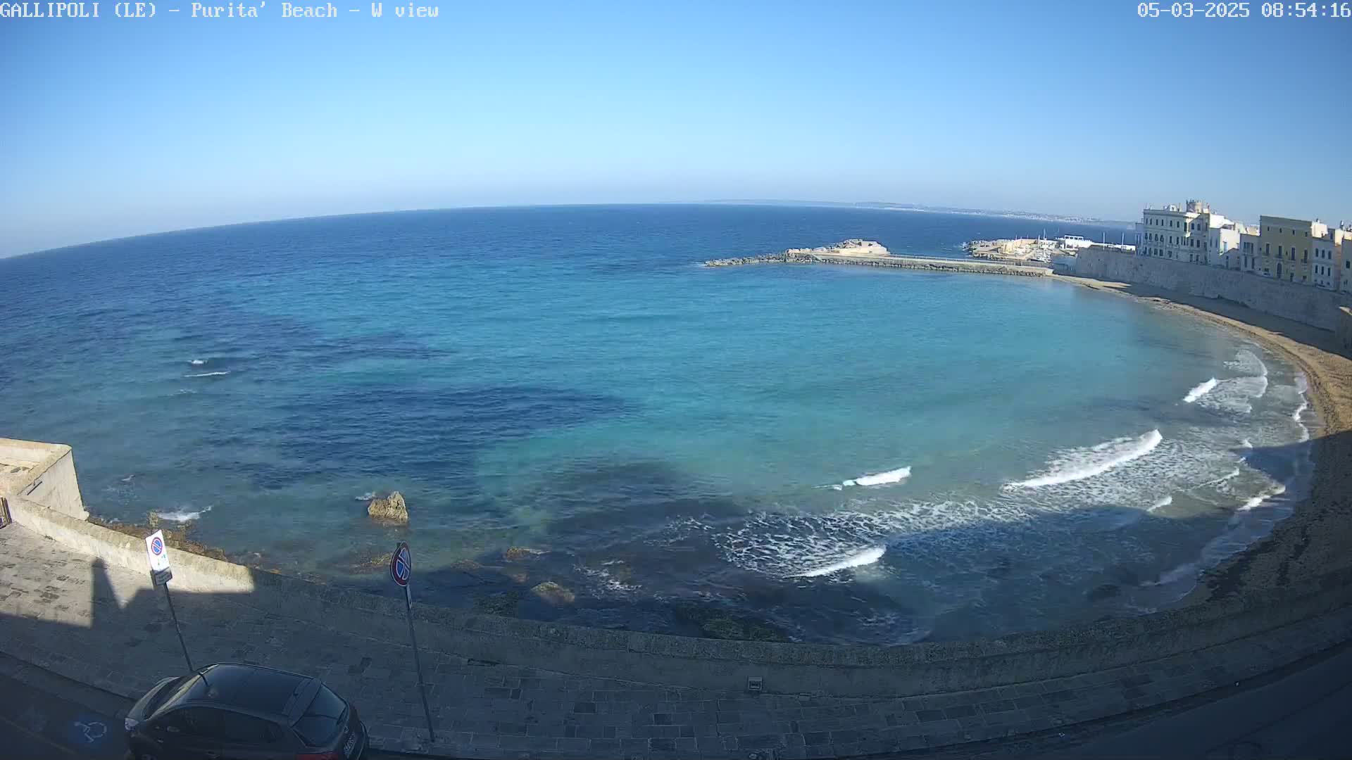 A clear, sunny day shows a curving, turquoise-blue bay with small waves lapping a sandy beach next to a stone-walled city.