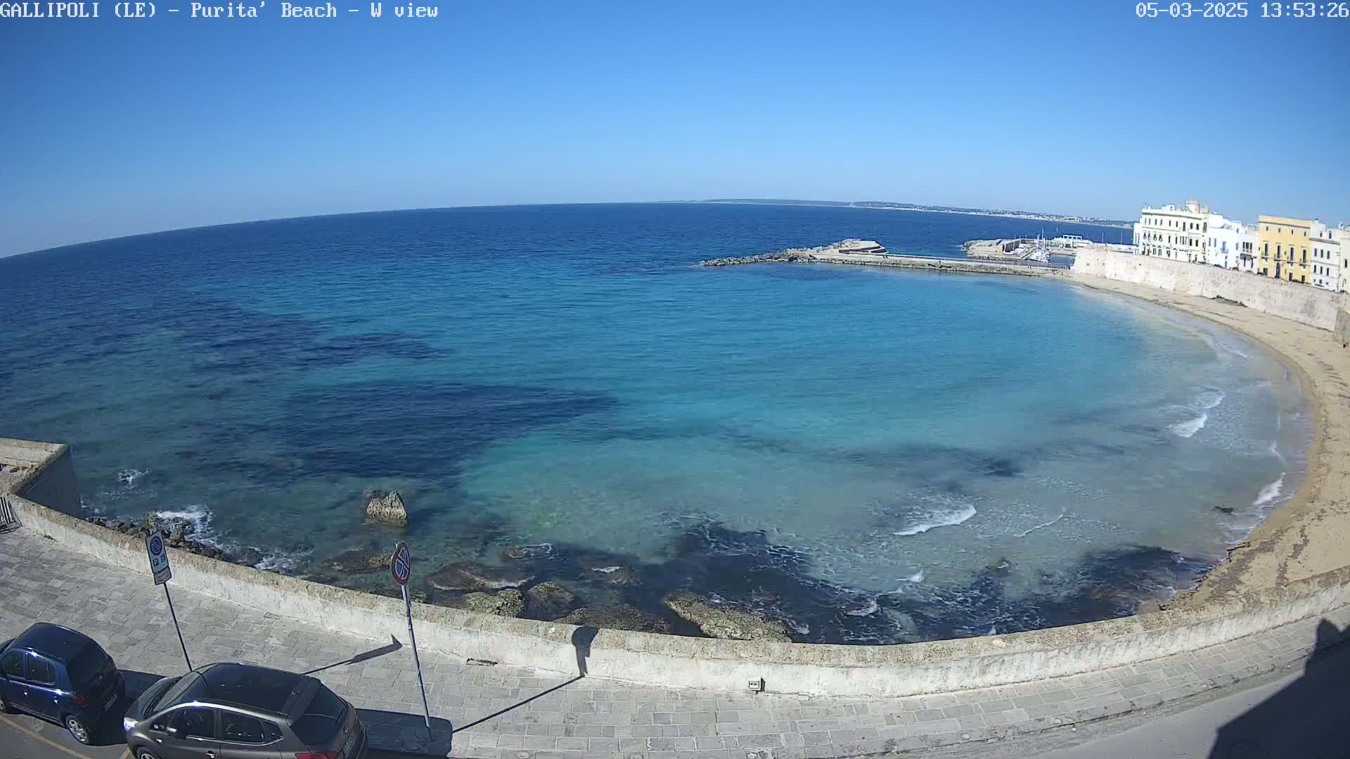 A clear, sunny day shows a curved beach with calm, shallow, turquoise water lapping against a stone wall, beside which two cars are parked.