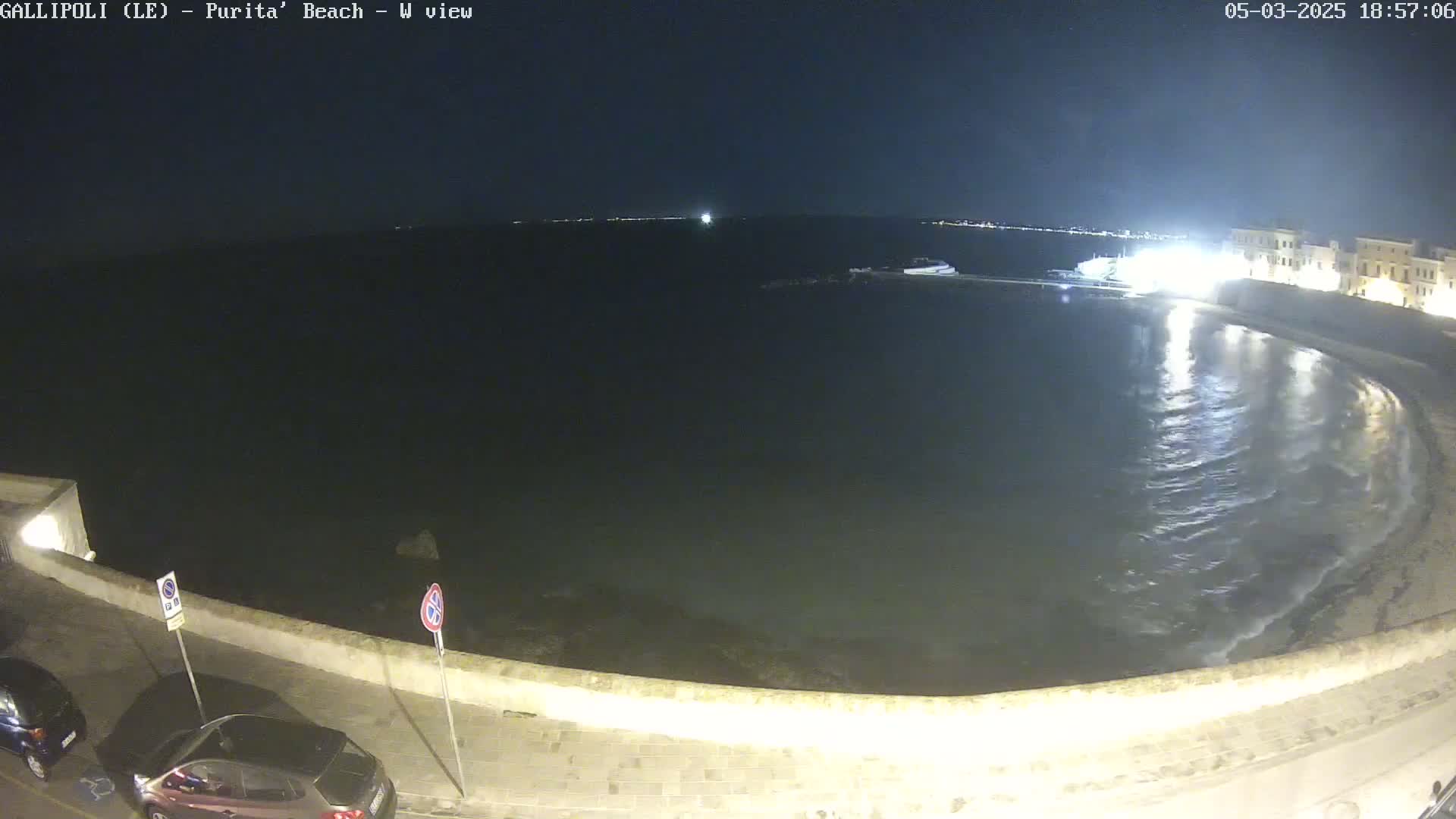 A nighttime view of a calm, dark sea lapping gently at a crescent-shaped beach next to a low stone wall and a street with parked cars.