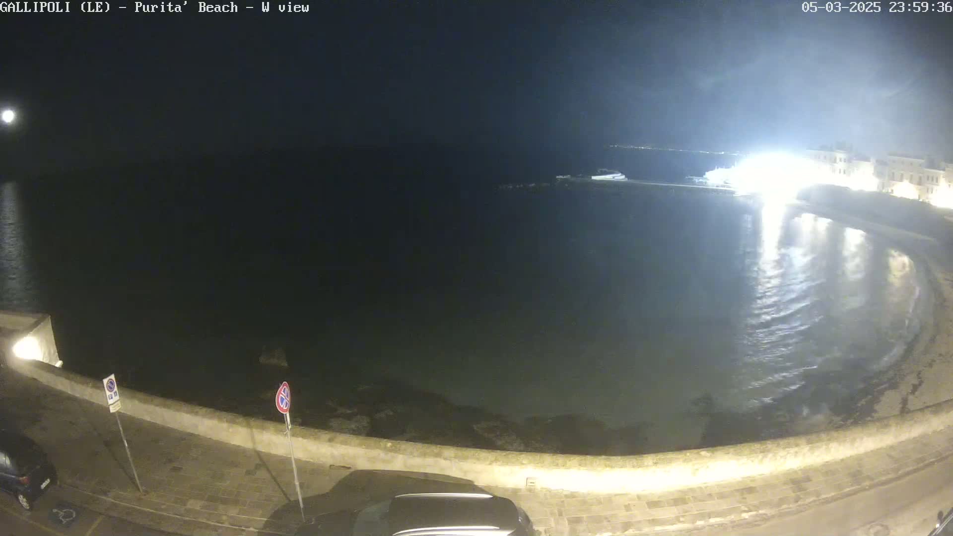 A nighttime view of a calm, dark body of water next to a stone wall, with a bright moon visible in the distance.
