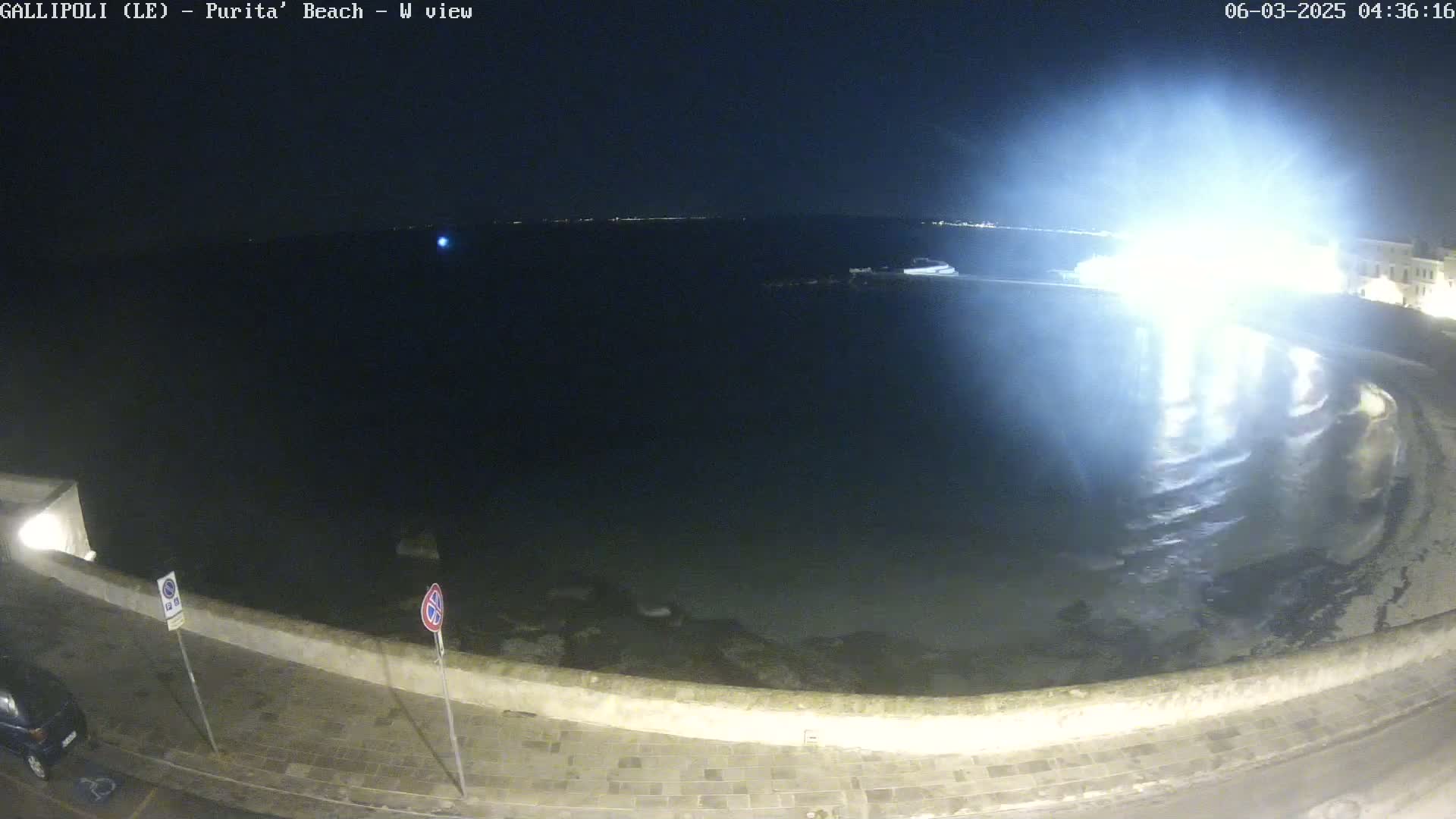 A nighttime view of a dark, calm body of water next to a paved shoreline, brightly lit by a nearby light source.