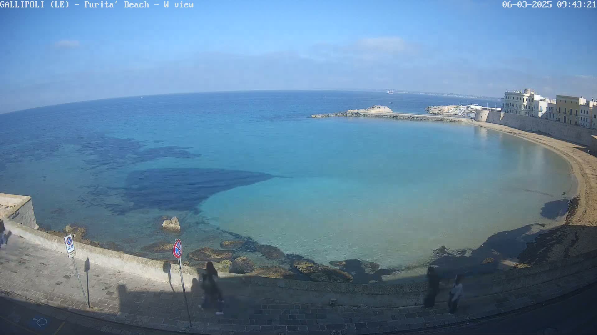 A clear, sunny day shows a calm, turquoise bay curving around a sandy beach and stone buildings.