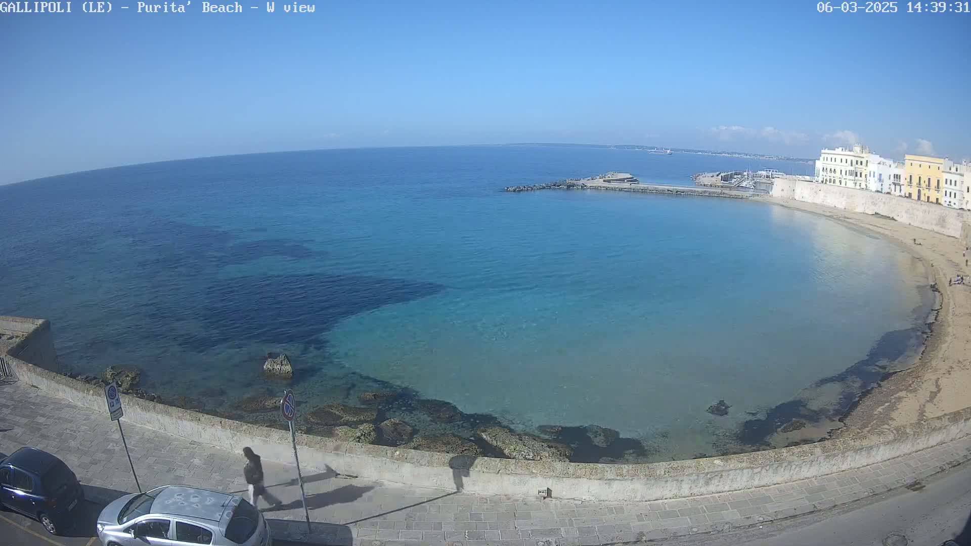 A clear, sunny day reveals a calm, turquoise bay with a sandy beach and buildings lining the shore, viewed from a high vantage point.
