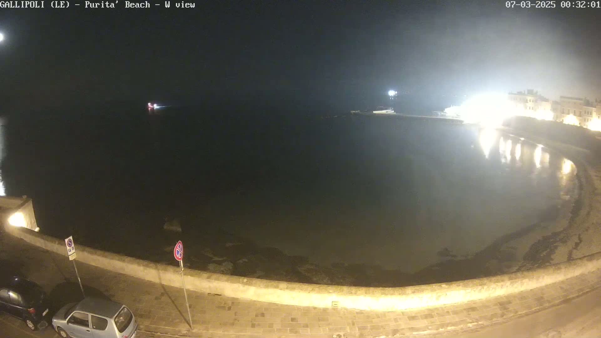 A nighttime, overhead view of a calm body of water next to a stone wall and paved area with two parked cars, under a mostly clear sky.