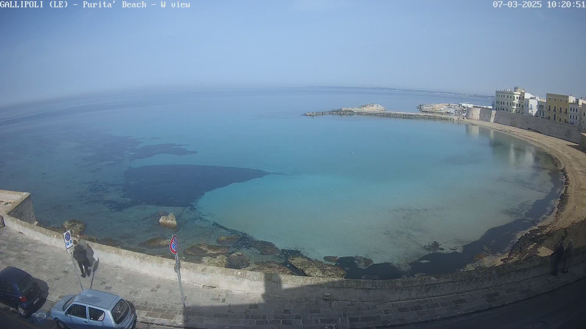 A clear, sunny day reveals a tranquil bay with calm, turquoise water lapping a sandy beach next to a stone wall and buildings.