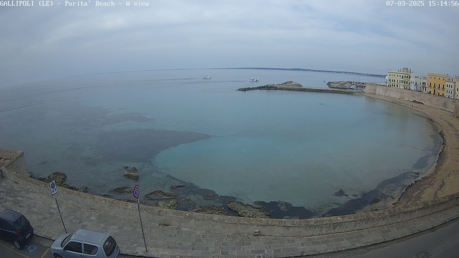 A partly cloudy day overlooks a calm, turquoise bay next to a sandy beach and stone buildings.