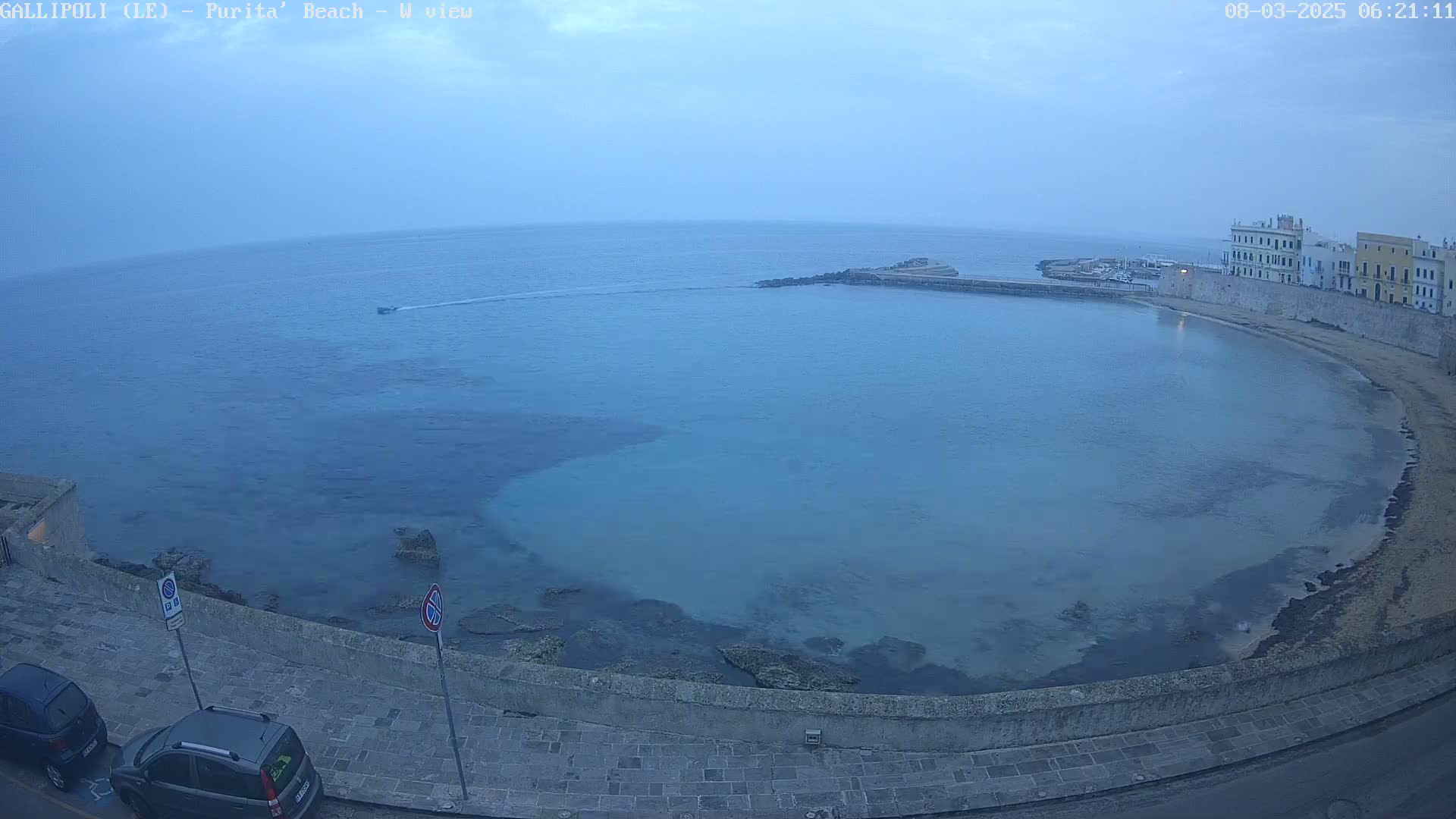 A calm, overcast day shows a bay with clear, shallow water and a small boat, next to a stone seawall and buildings.