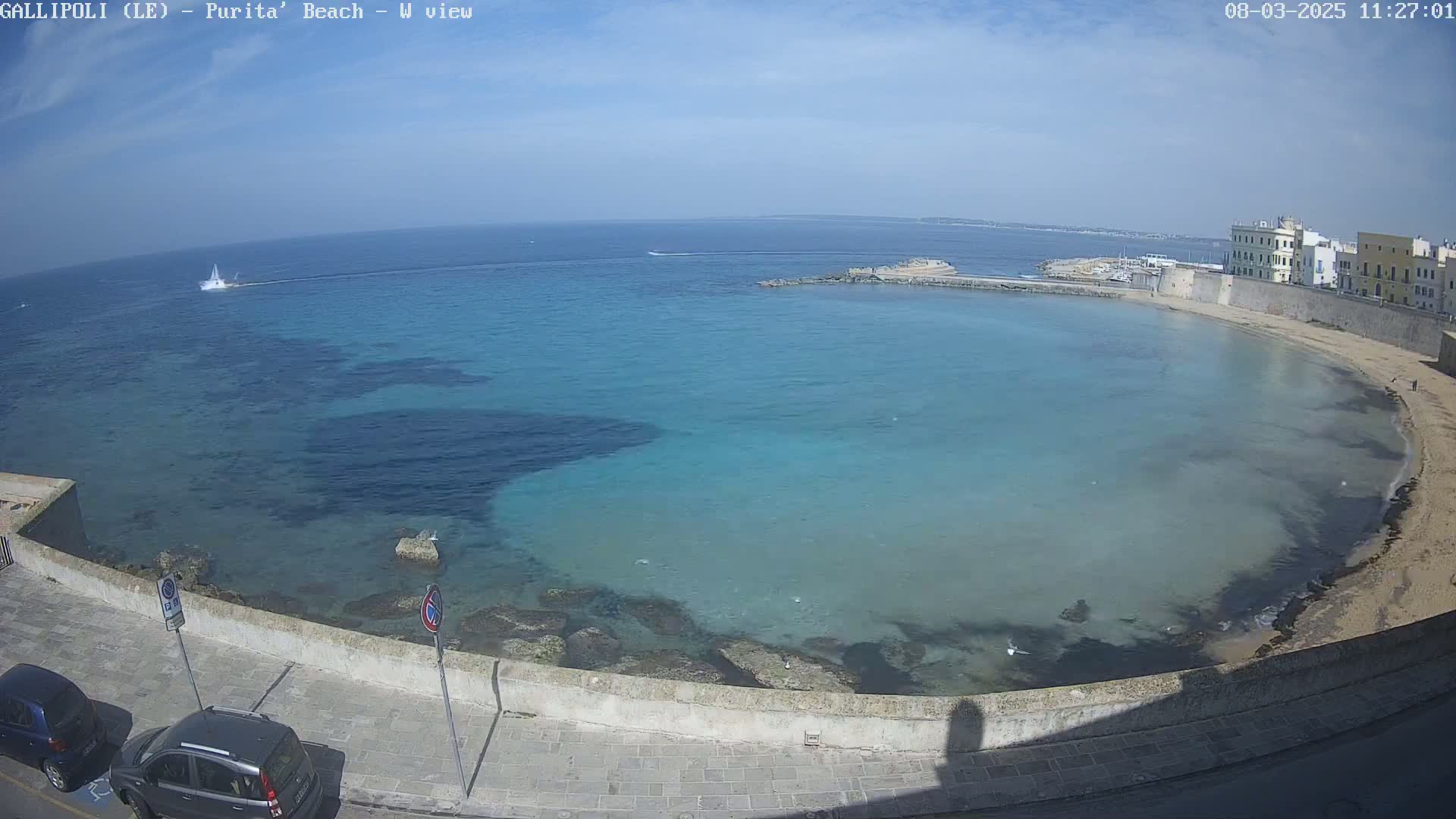 A clear, sunny day overlooks a calm, turquoise bay with a sandy beach and buildings along the shoreline.