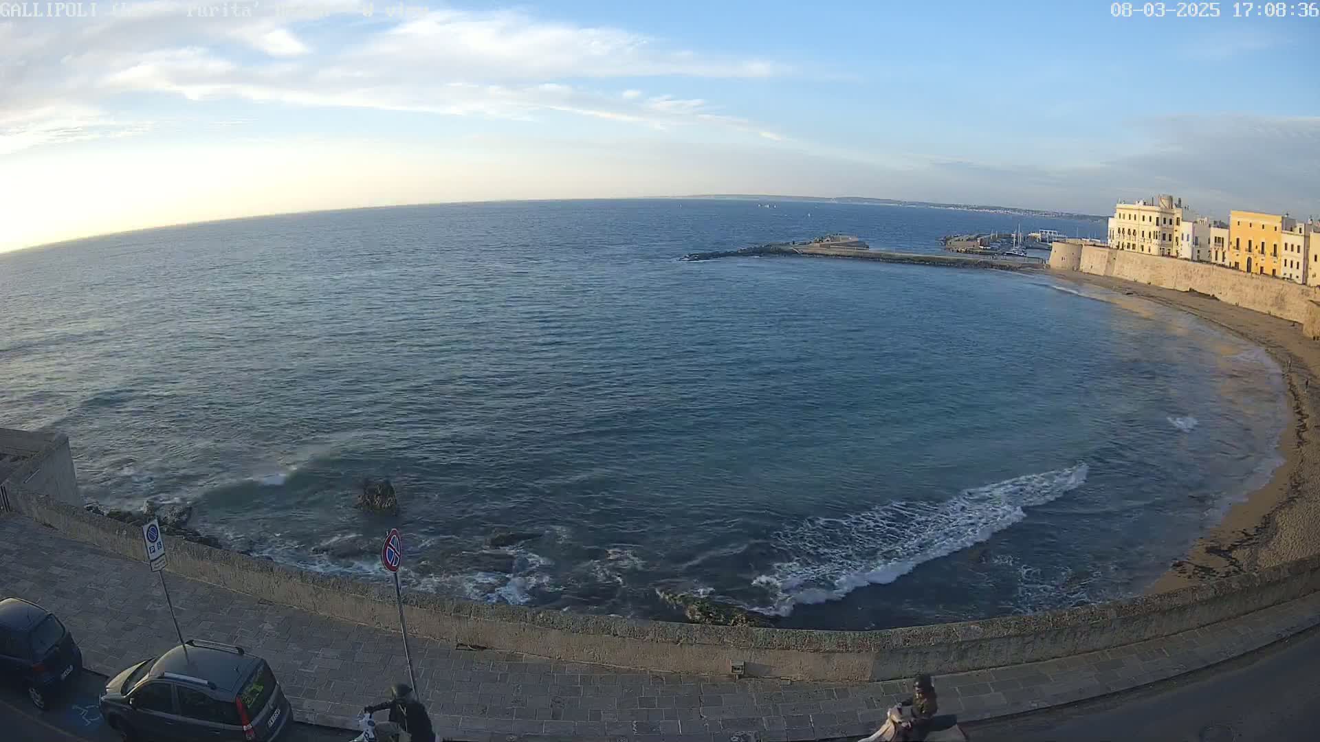 A partly sunny day shows a calm sea lapping a sandy beach beside a stone wall and buildings.
