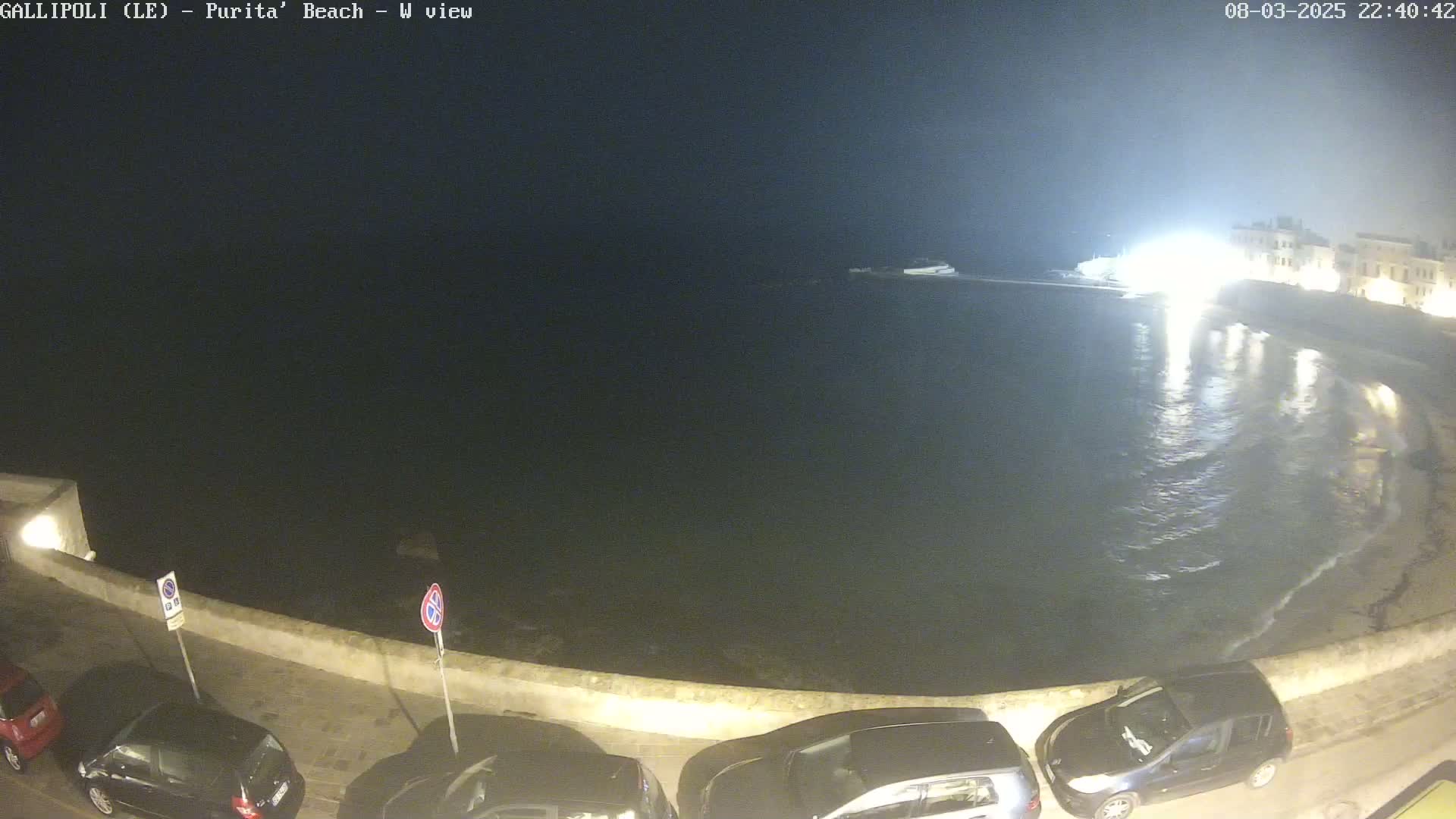 A nighttime view of a calm, dark body of water next to a seawall with several cars parked along it.
