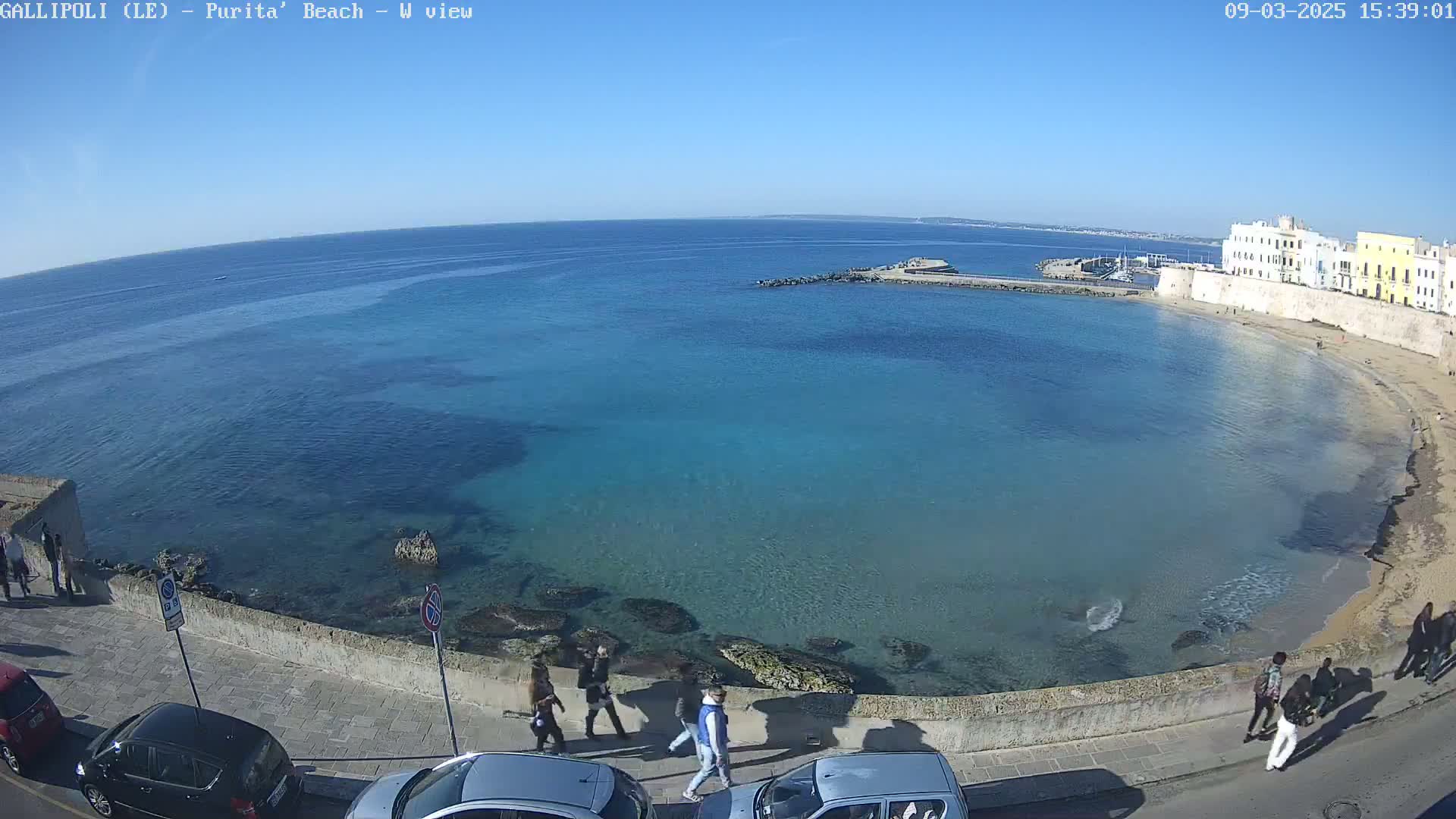 A clear, sunny day at a beach with turquoise water, a stone wall, parked cars, and several people walking along a paved path.