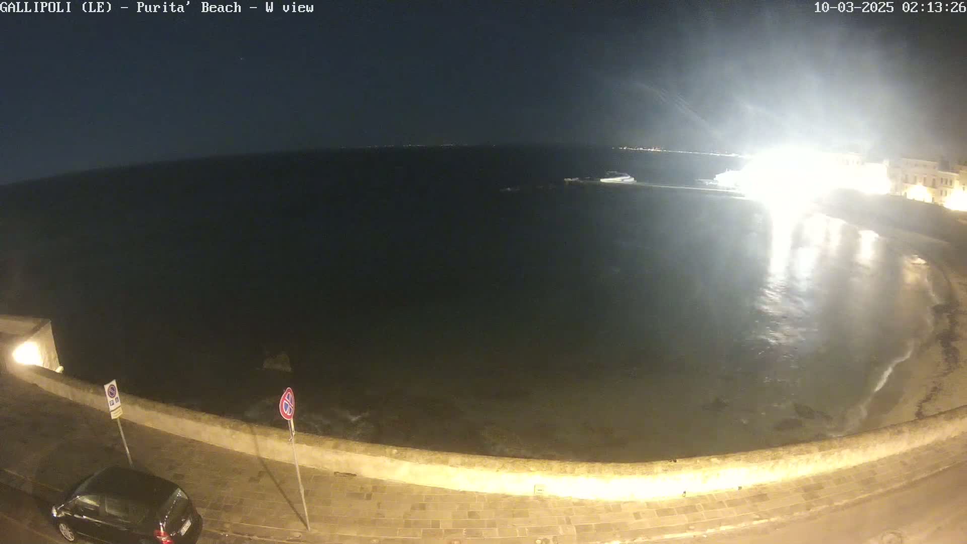 A nighttime view of a calm sea lapping a stone wall next to a paved road, with a car parked nearby and brightly lit buildings in the distance.