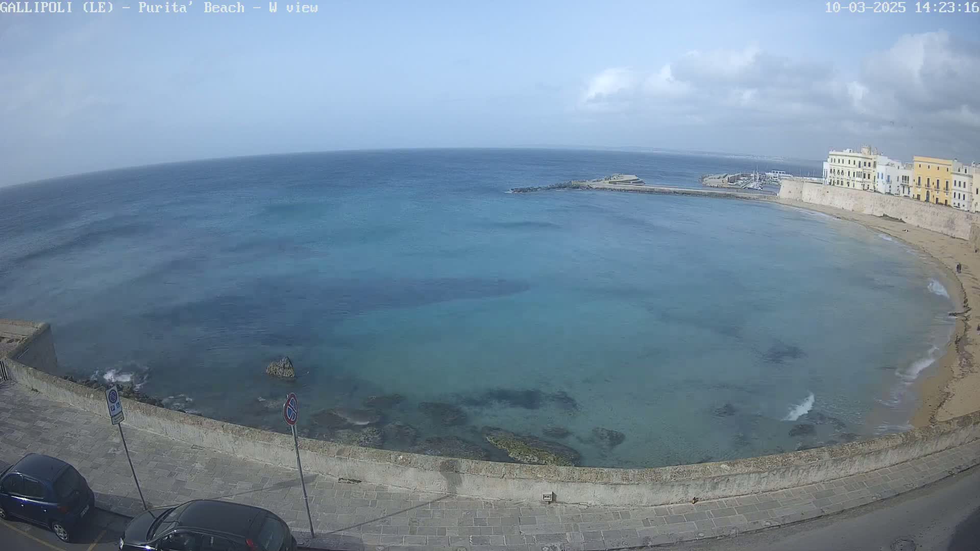 A partly cloudy day reveals a calm, clear turquoise bay curving around a sandy beach and stone buildings.