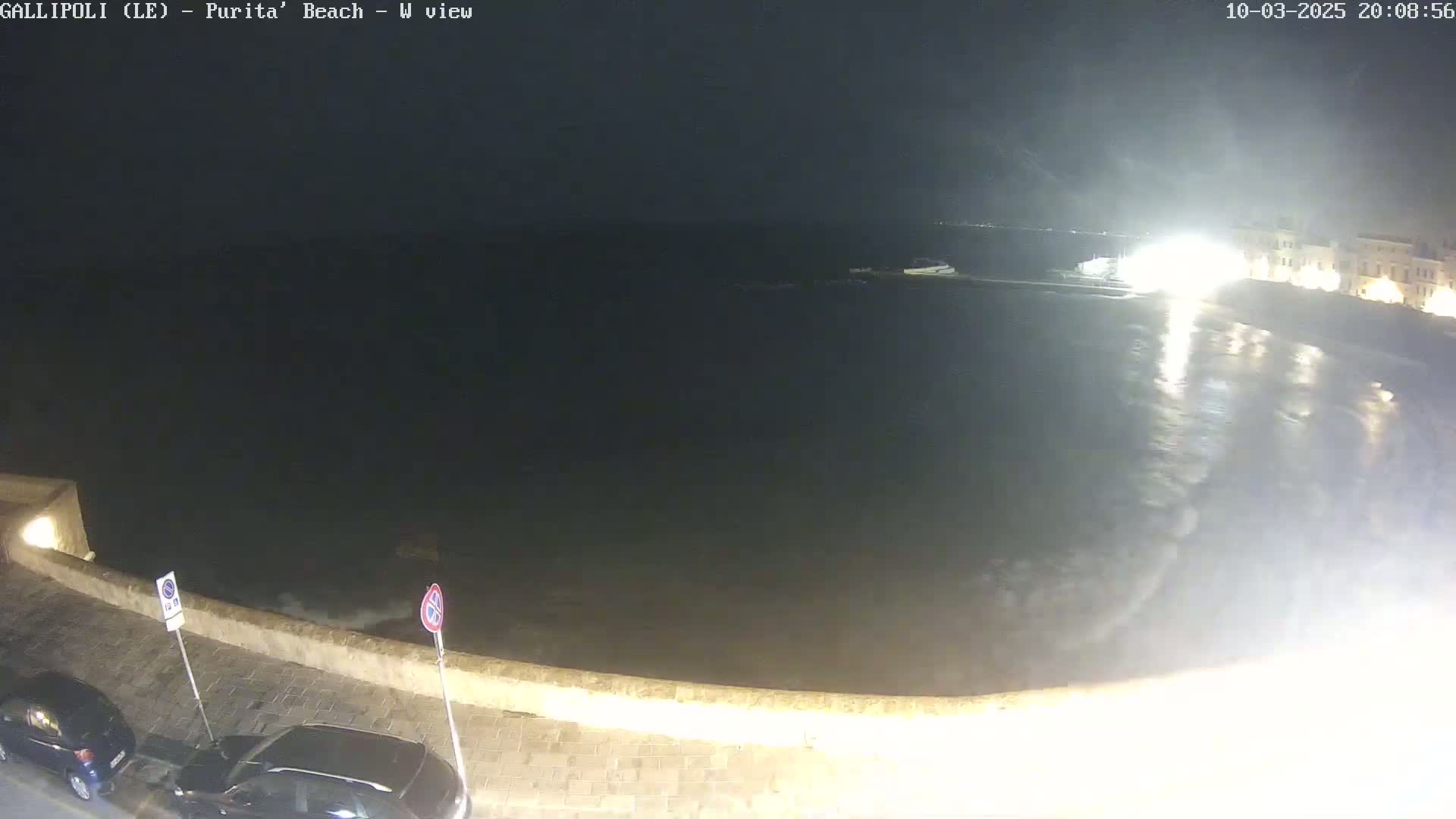 A nighttime view of a dark, wavy ocean next to a stone wall with parked cars, under a somewhat overcast sky.