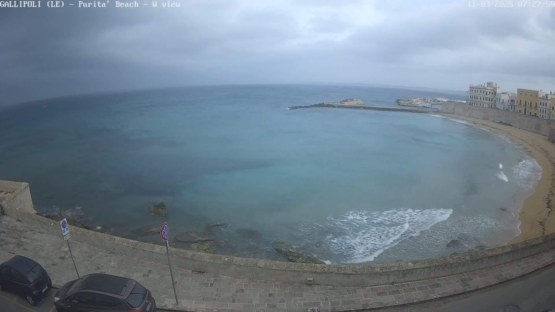 A partly cloudy day shows a view of a calm, teal bay with small waves lapping a sandy beach next to a stone wall and buildings.