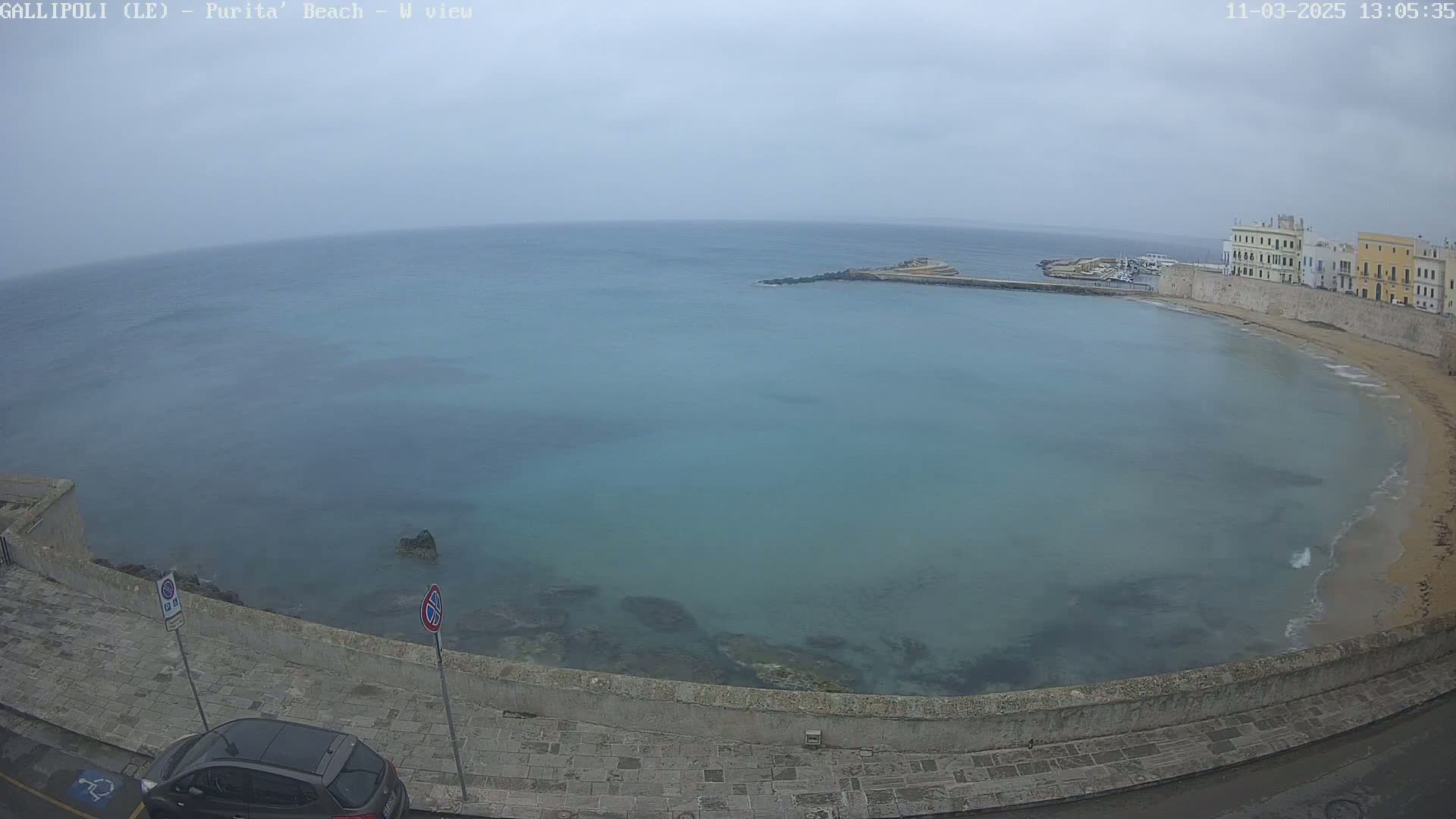 A gray, overcast day shows a calm, teal-colored bay curving around a stone embankment, where a small car is parked near buildings in the background.