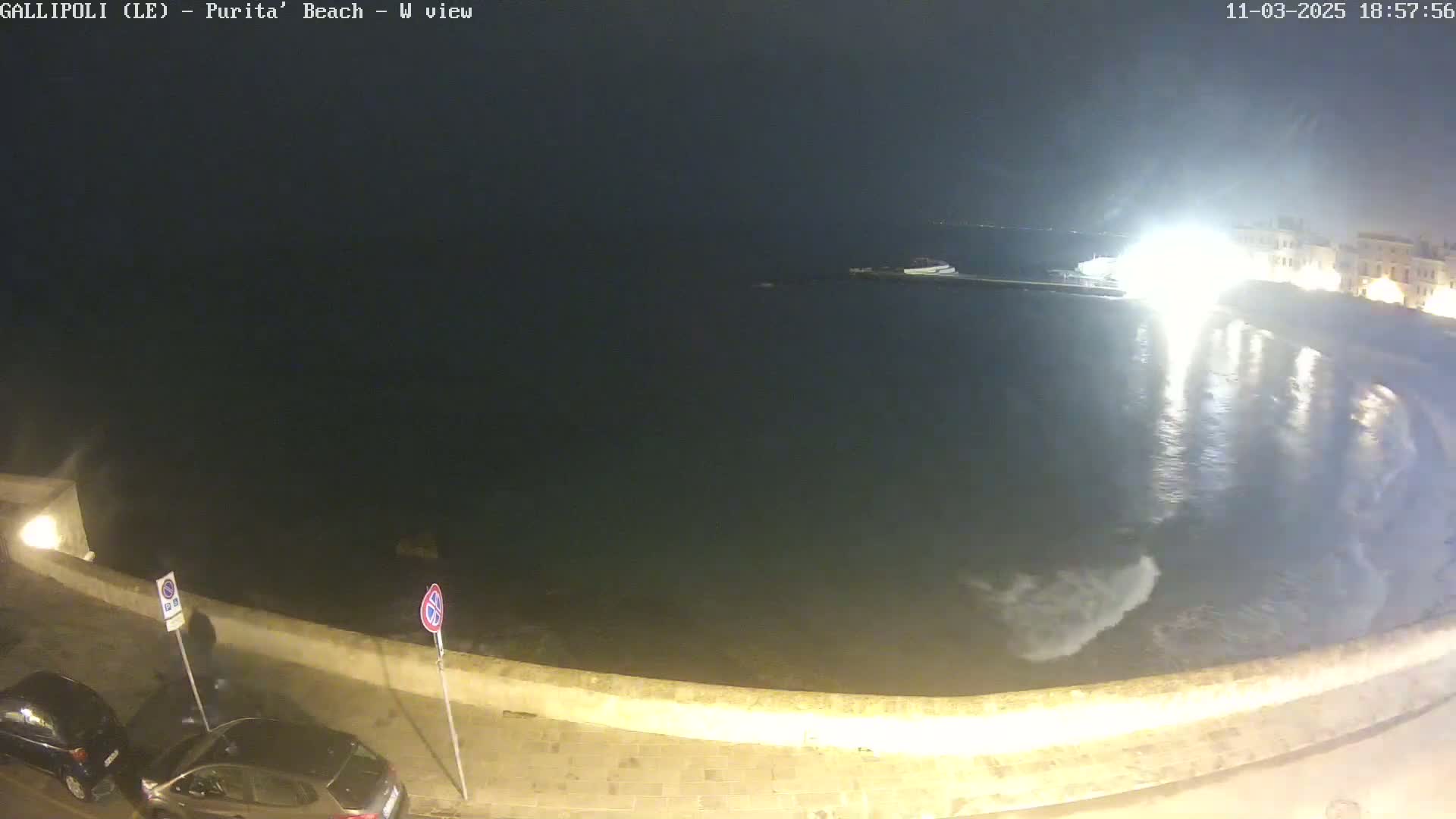 At night, a dark body of water laps gently against a low stone wall, next to which two cars are parked.