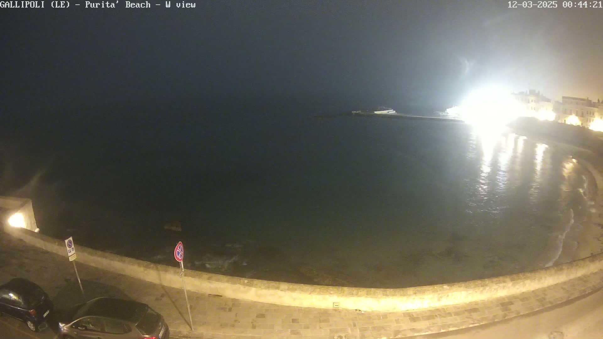 A nighttime view of a calm sea next to a stone wall and roadway, with a brightly lit building in the background under a hazy sky.