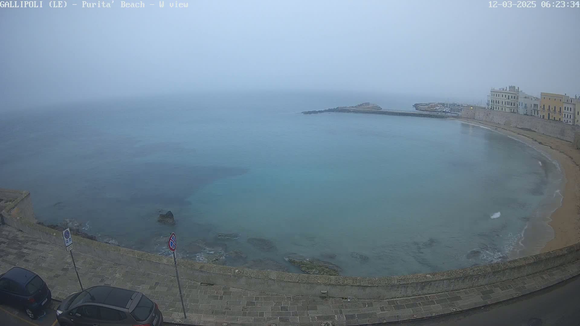 A foggy view of a calm, turquoise bay curving along a stone seawall with buildings and parked cars visible along the shore.