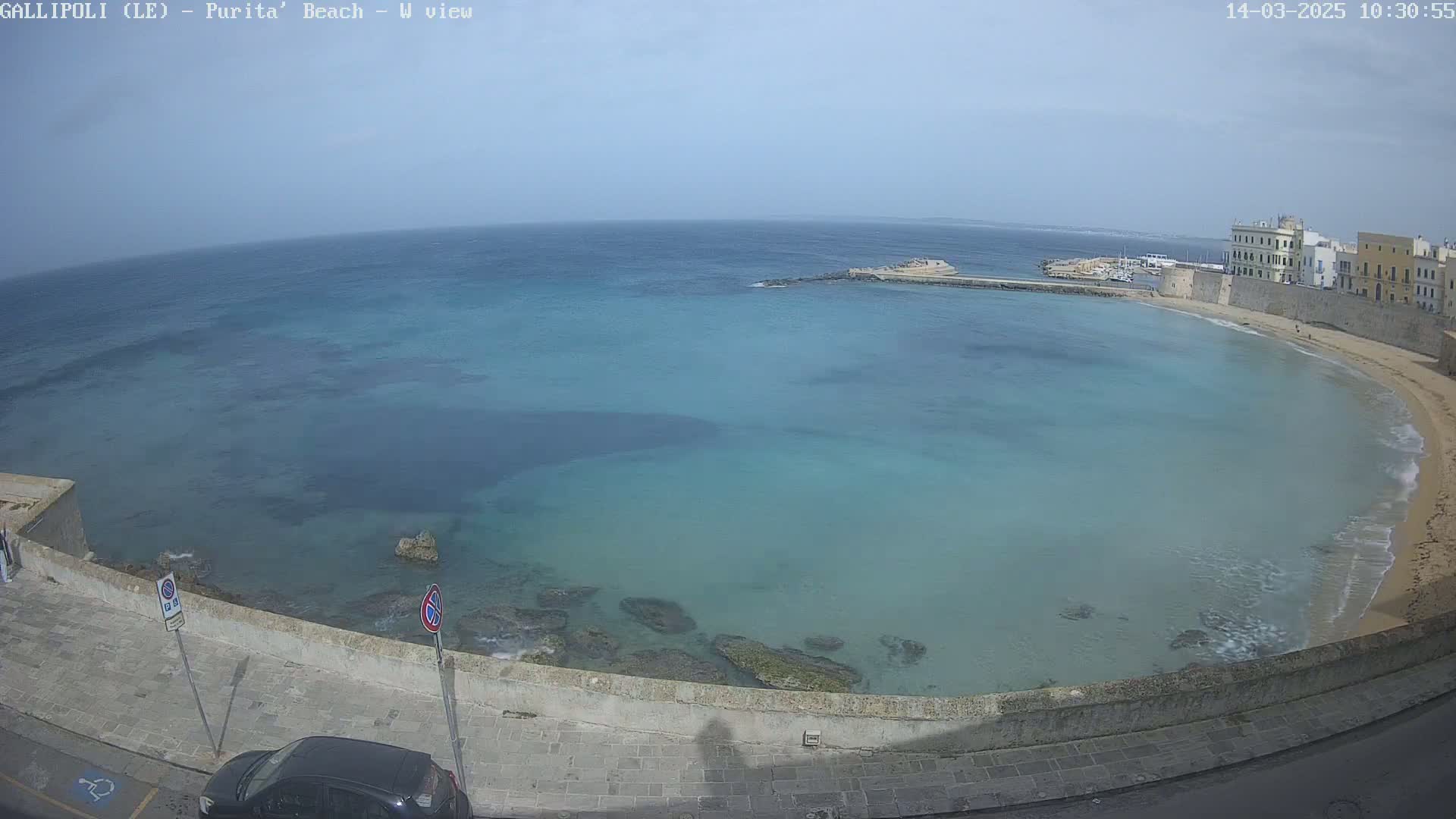 A clear, sunny day overlooks a calm, turquoise bay with a sandy beach curving around a stone wall and buildings.