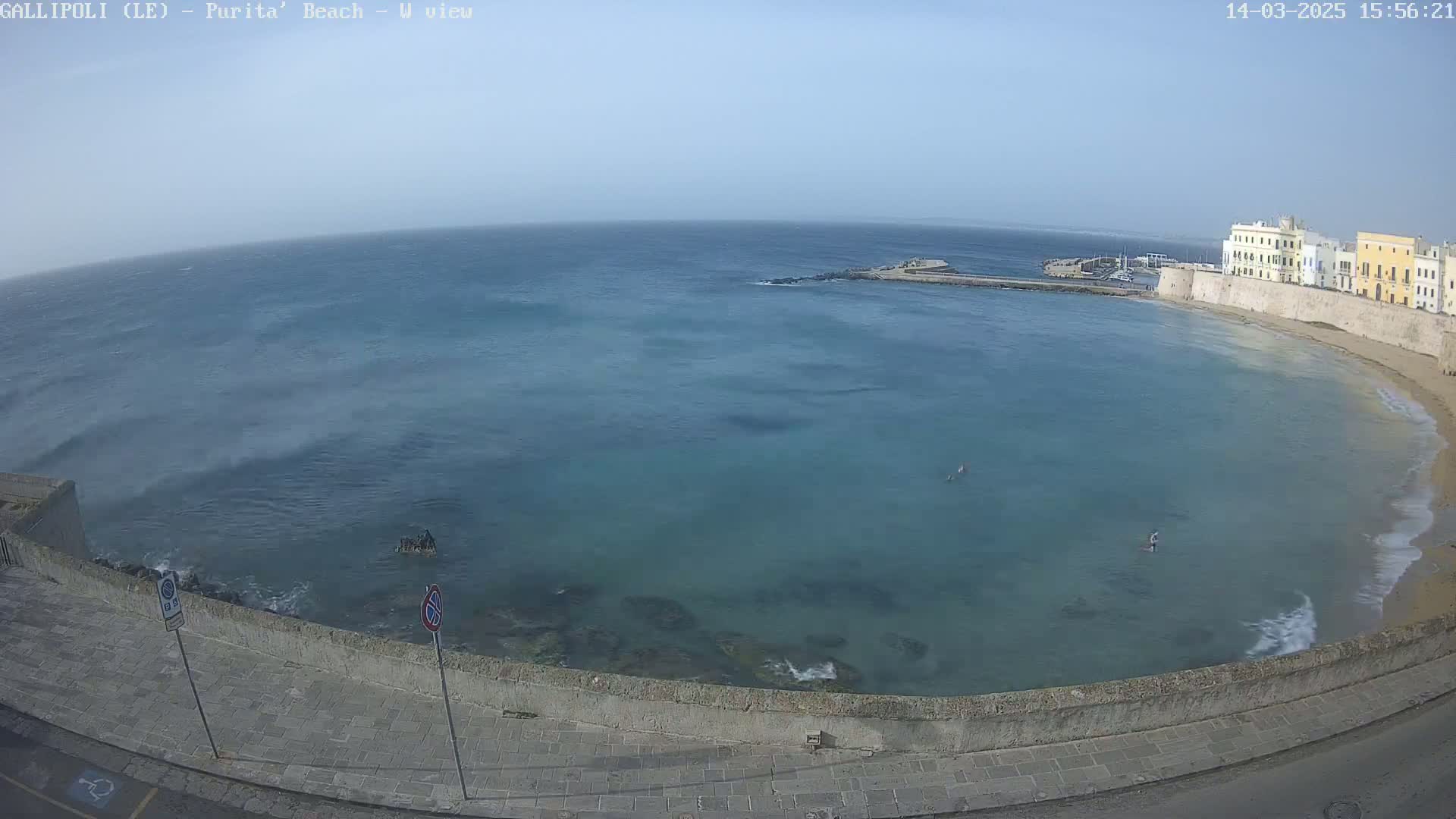 A partly sunny day shows a calm sea curving around a stone wall and buildings, with a few people in the water.