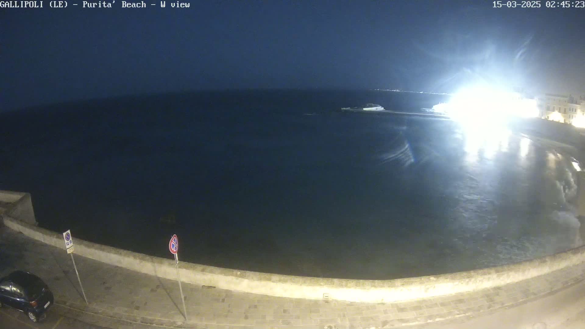 A nighttime, wide shot of a calm, dark sea lapping against a low stone wall next to a paved road with a car parked.