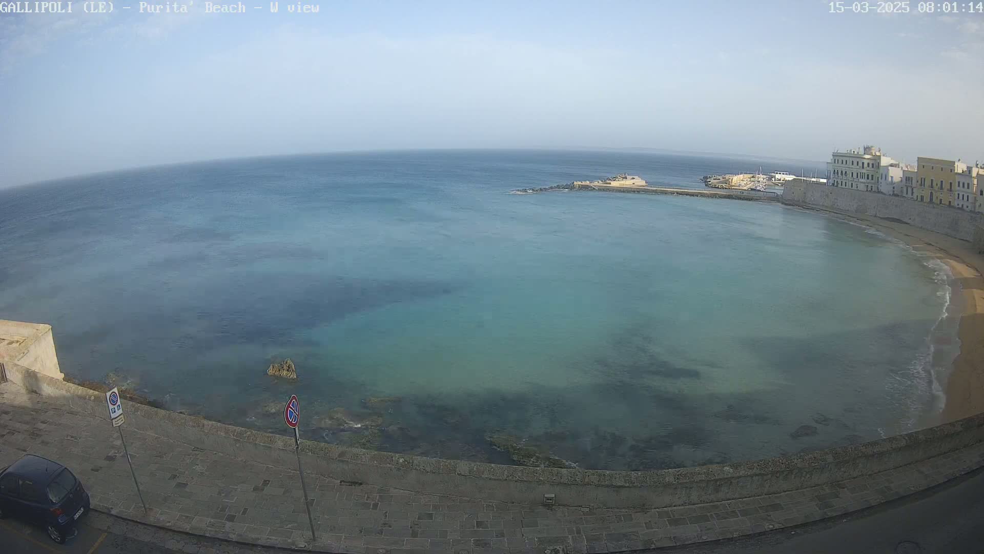 A calm, clear day shows a tranquil bay with turquoise water lapping a sandy beach, bordered by stone walls and buildings.