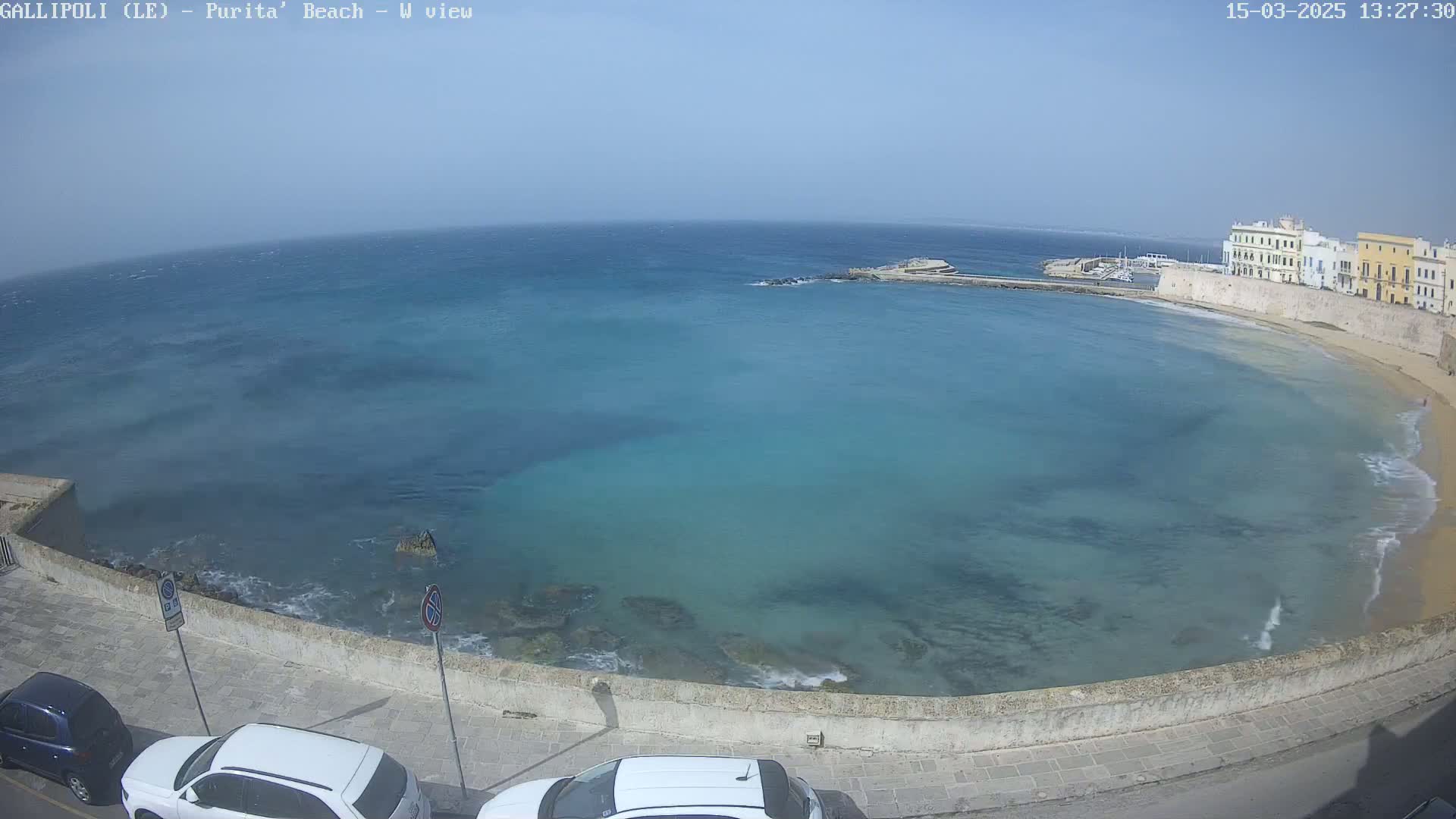 A clear, sunny day overlooks a calm, turquoise bay with a sandy beach and buildings along the shoreline.
