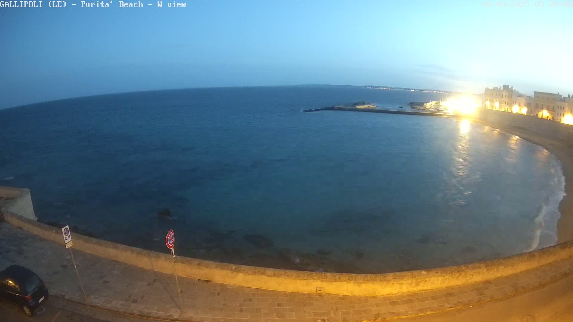 A calm, dark-blue sea laps gently against a low stone wall at dusk, with a town's illuminated buildings visible in the background.