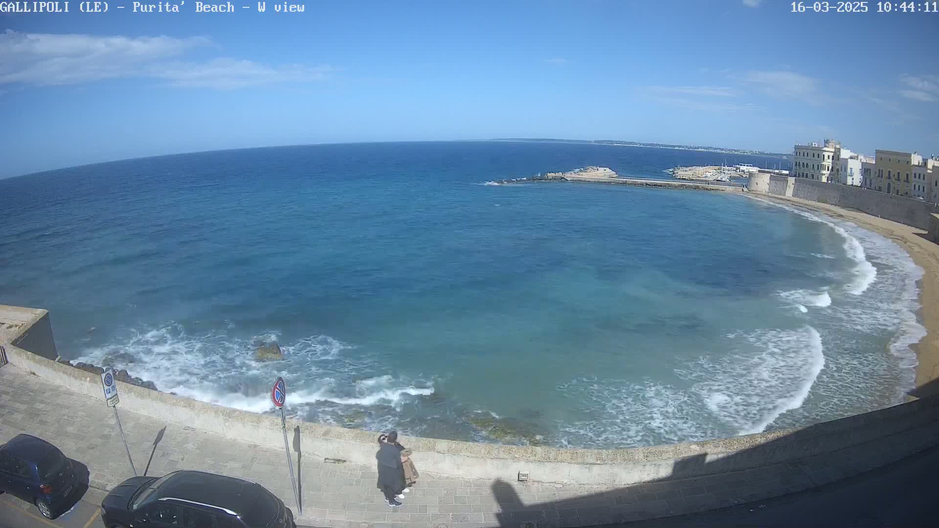 A clear, sunny day shows a coastal view of a calm, blue sea with small waves lapping a sandy beach, backed by a stone wall and buildings.