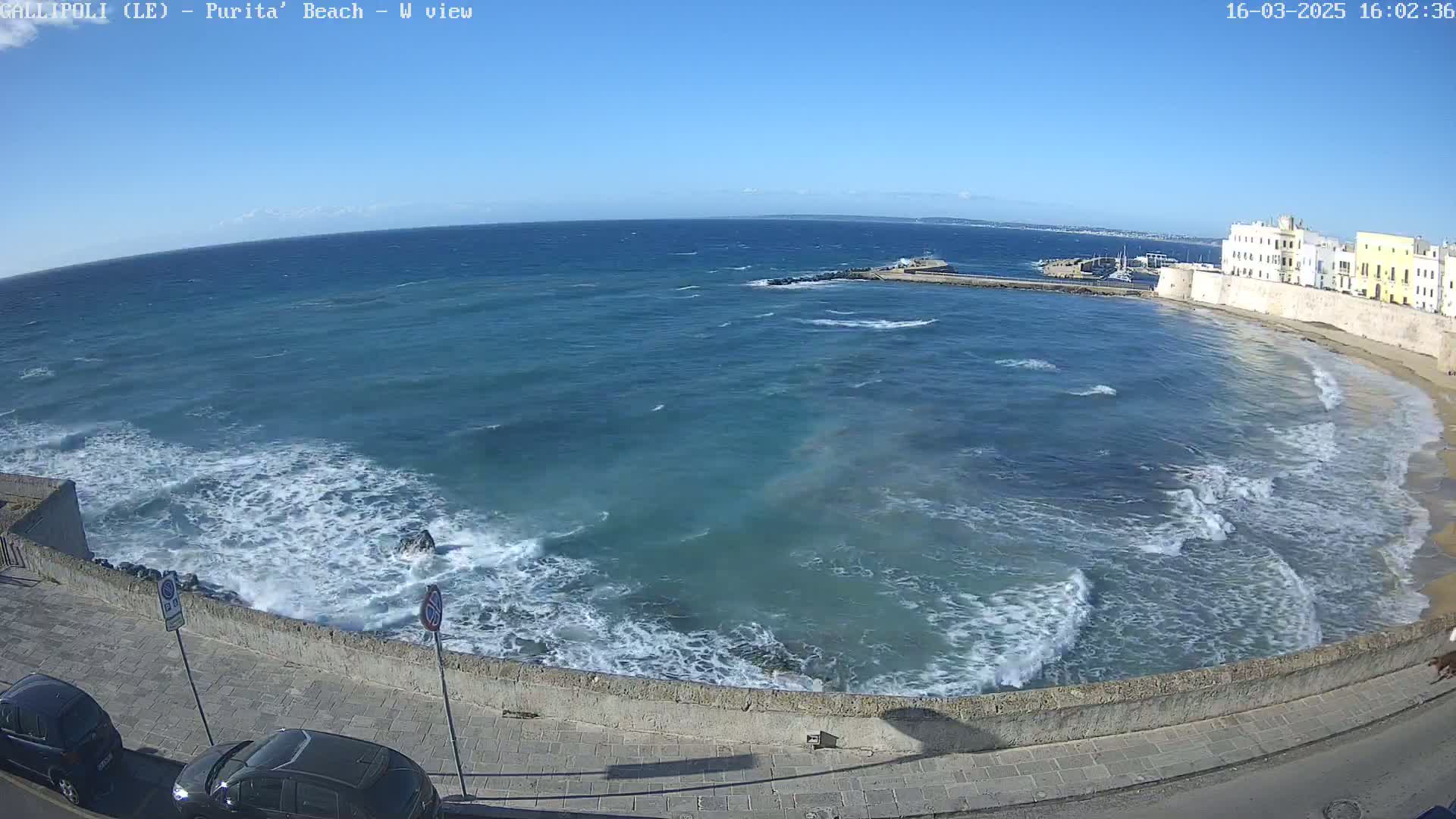 A beach with moderate waves under a clear blue sky is viewed from above, showing a stone wall and parked cars along the shore.