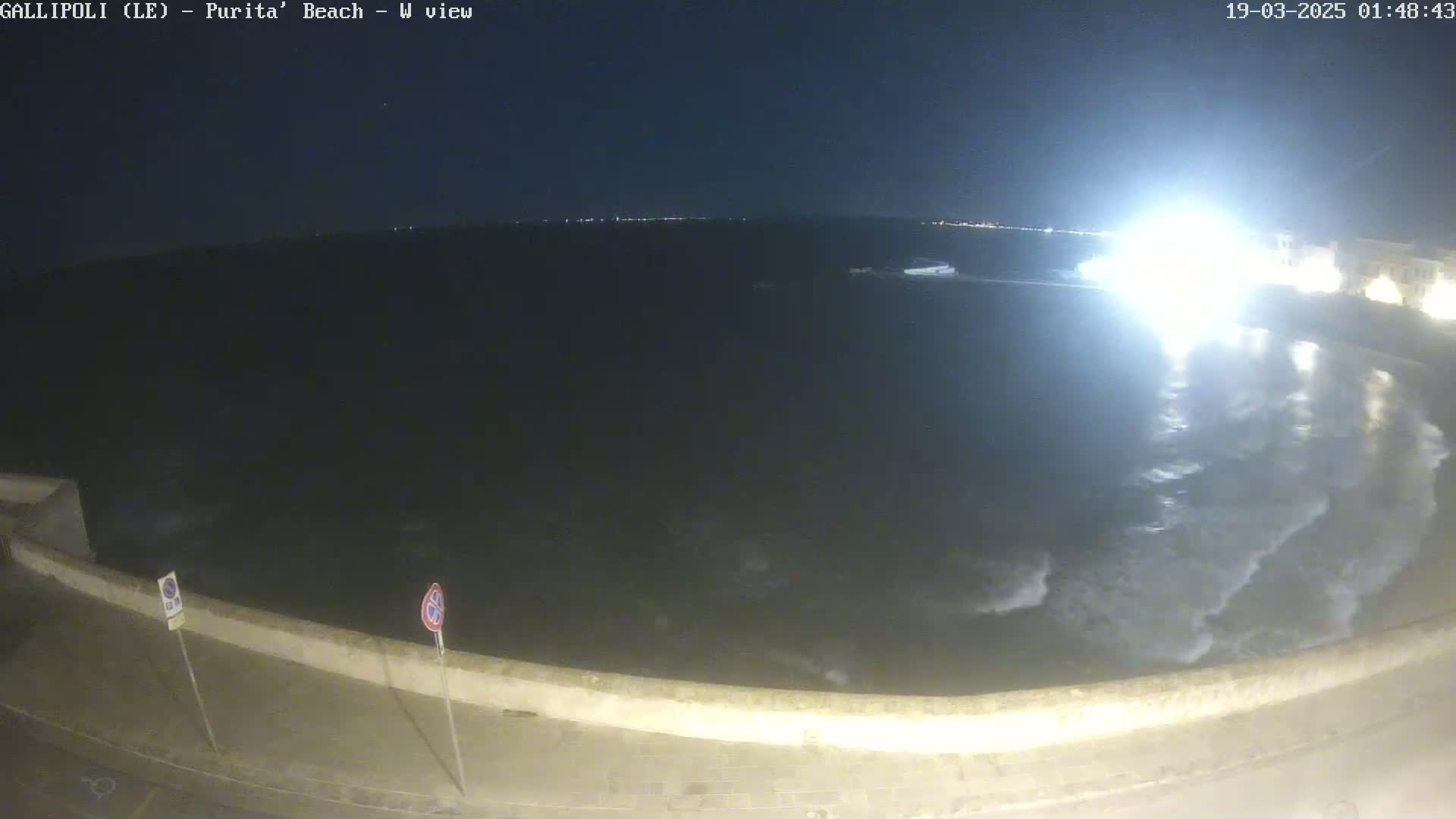 A nighttime, wide shot of a dark sea with small waves lapping against a low wall next to a paved road.