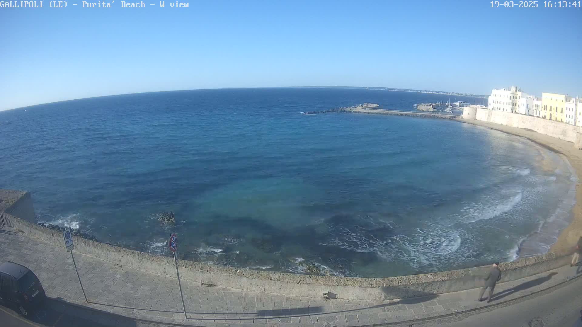 A clear, sunny day shows a calm, blue sea lapping a sandy beach next to a stone wall and buildings.