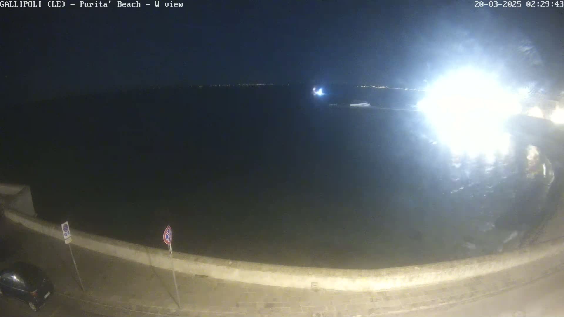 A nighttime view of a dark body of water, a low wall, and a parked car under a clear sky.