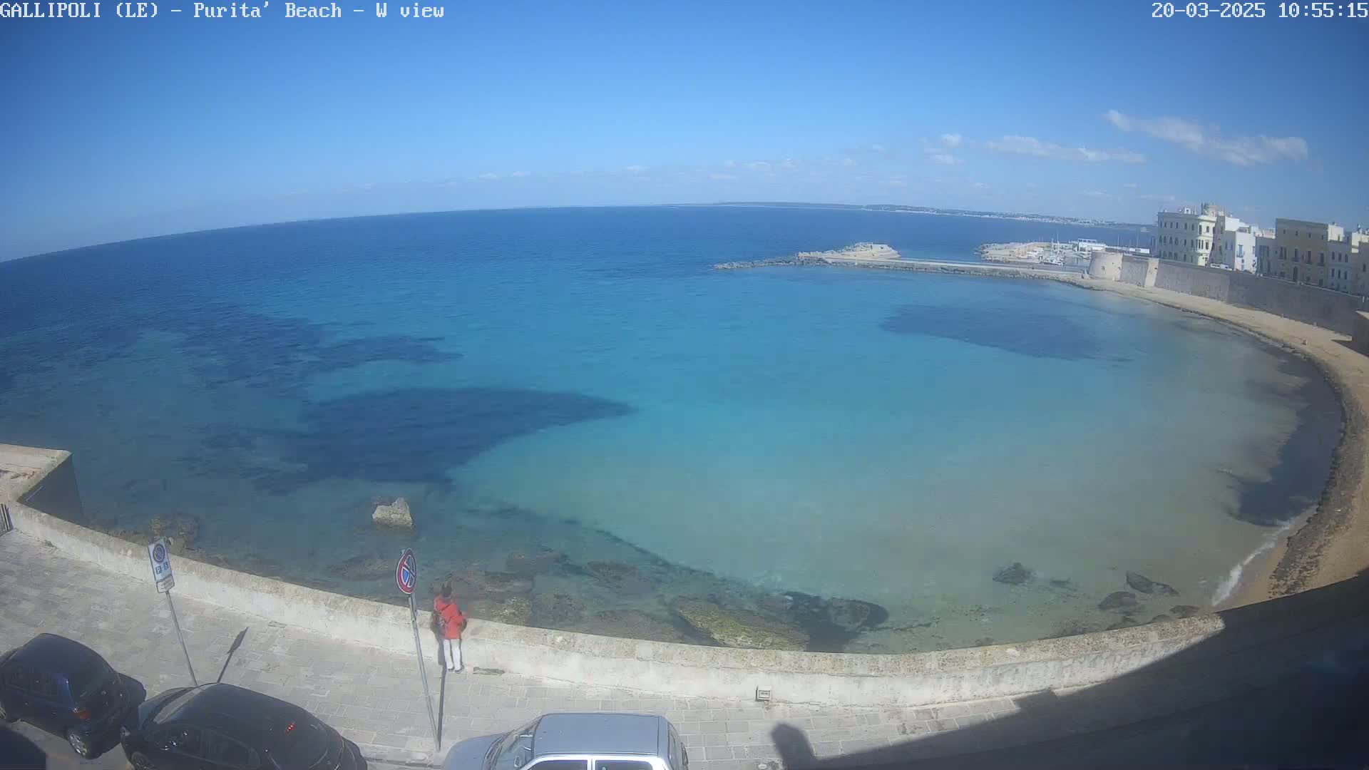 A high-angle view of a calm, clear turquoise sea curving around a sandy beach and stone wall under a mostly sunny sky.