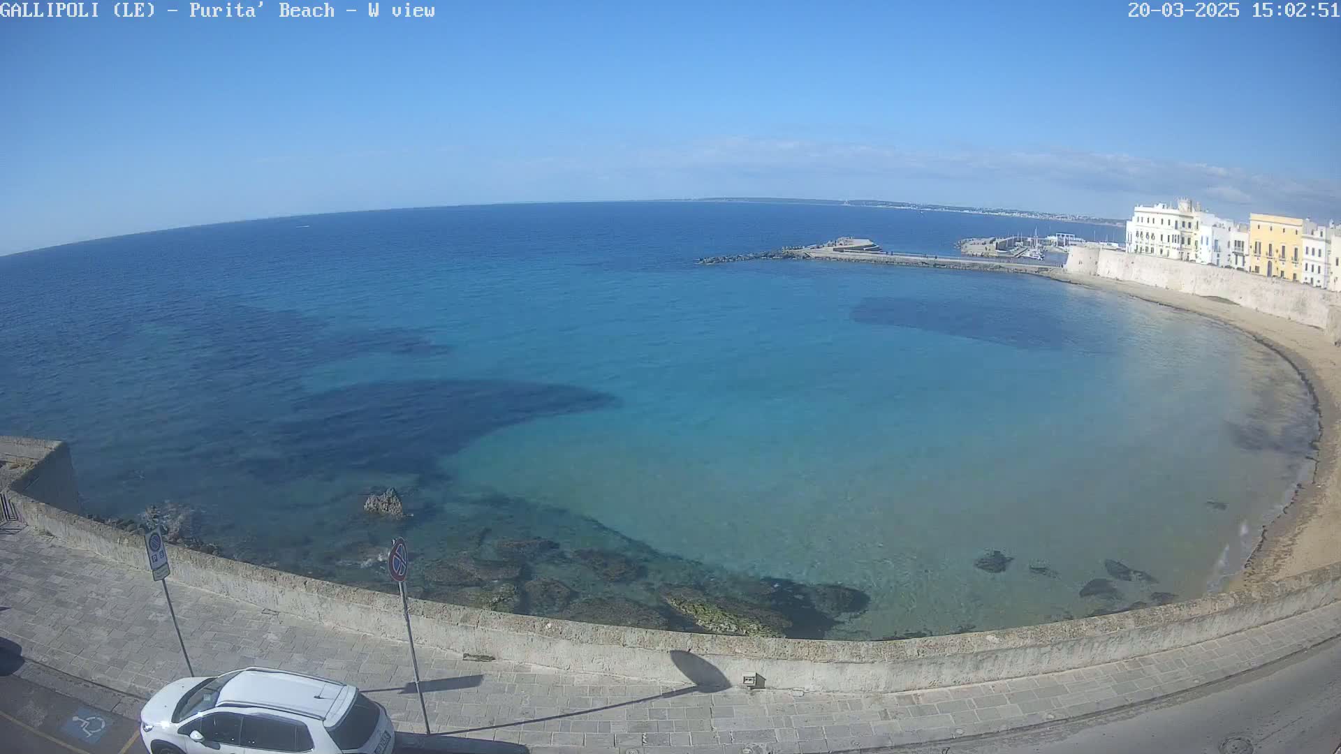 A clear, sunny day overlooks a calm, turquoise bay with a sandy beach and buildings along the shore.