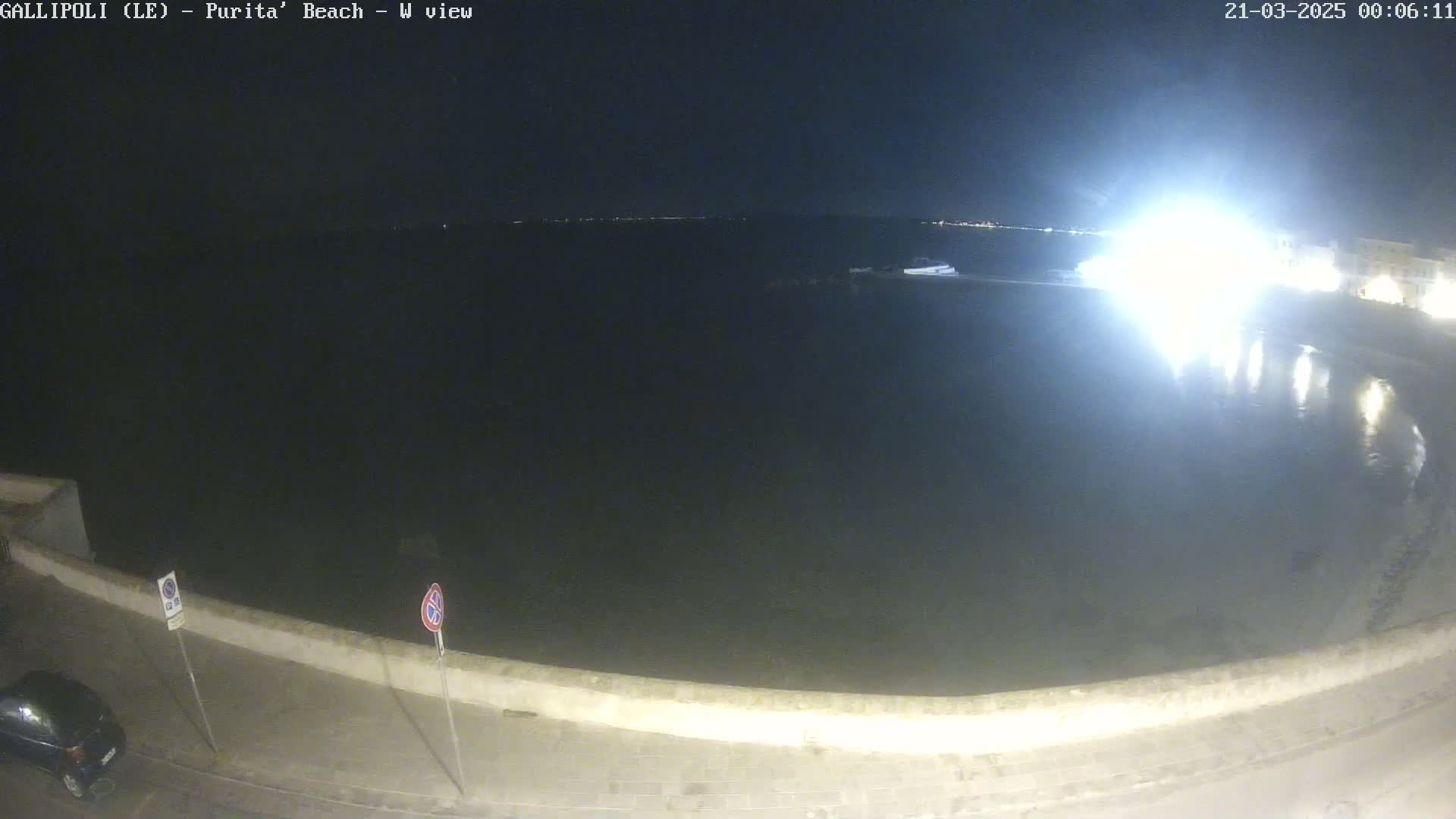 A nighttime view of a calm body of water next to a low wall, with a car parked on the road beyond.