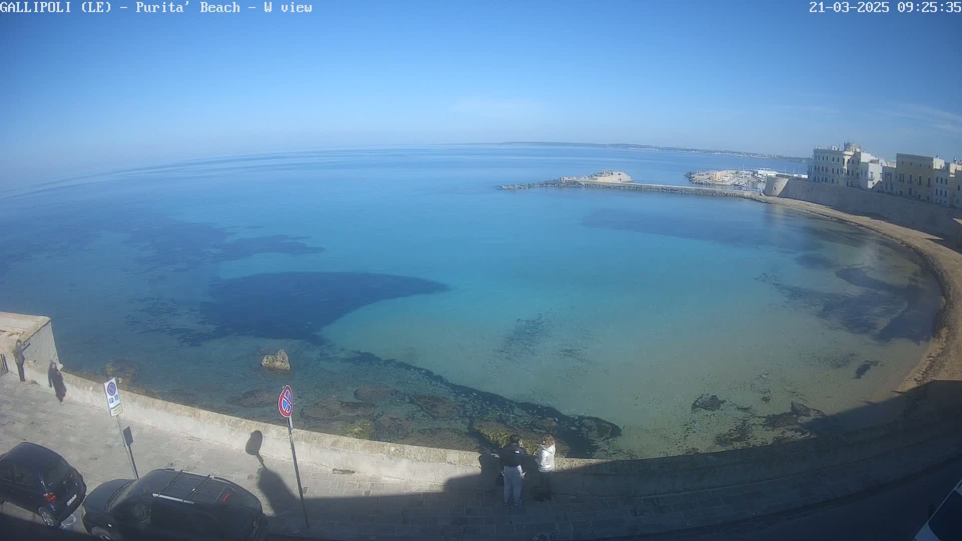 A clear, sunny day reveals a calm, turquoise-blue bay curving along a stone wall, with buildings visible in the distance and two people standing near the water's edge.