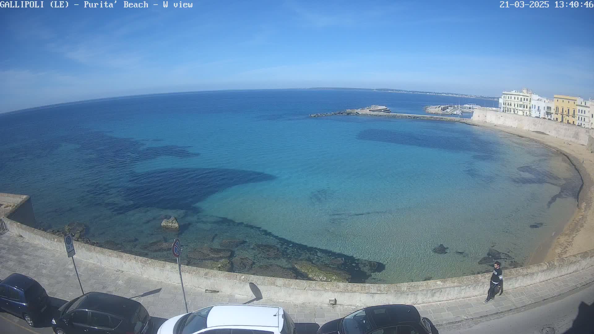 A clear, sunny day reveals a picturesque coastal scene with turquoise water lapping a sandy beach next to a stone wall and buildings, and several cars parked nearby.