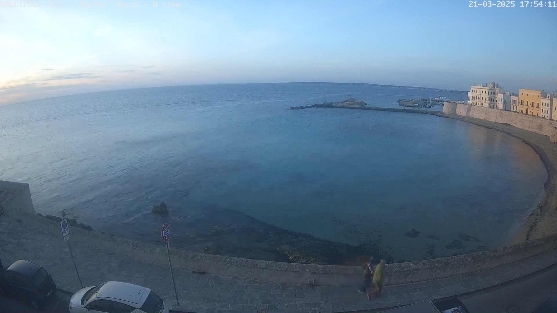 A high-angle, long shot shows a calm, clear body of water curving around a stone-walled city under a clear, twilight sky; two people walk along a paved path next to the water.