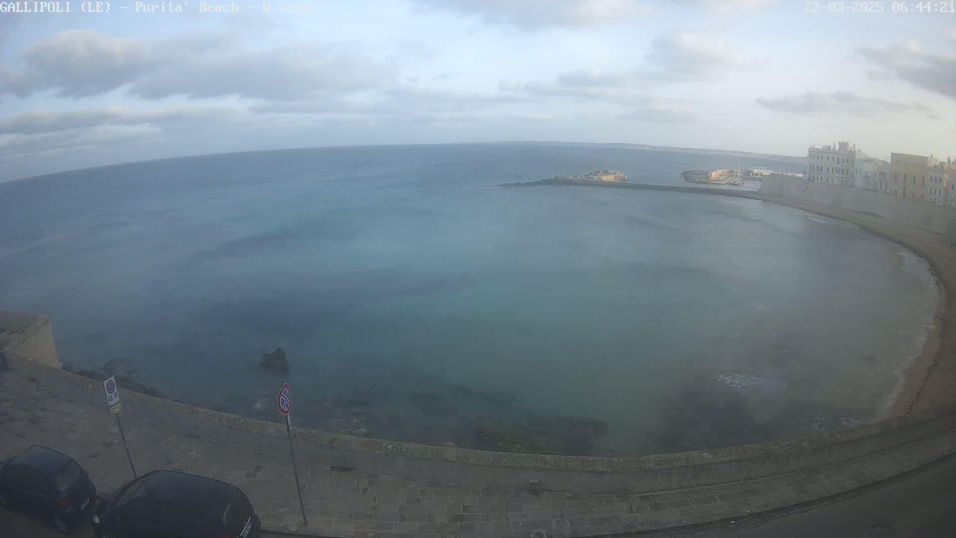 A partly cloudy day reveals a calm, teal-green sea curving into a sandy shore next to a stone wall and buildings.