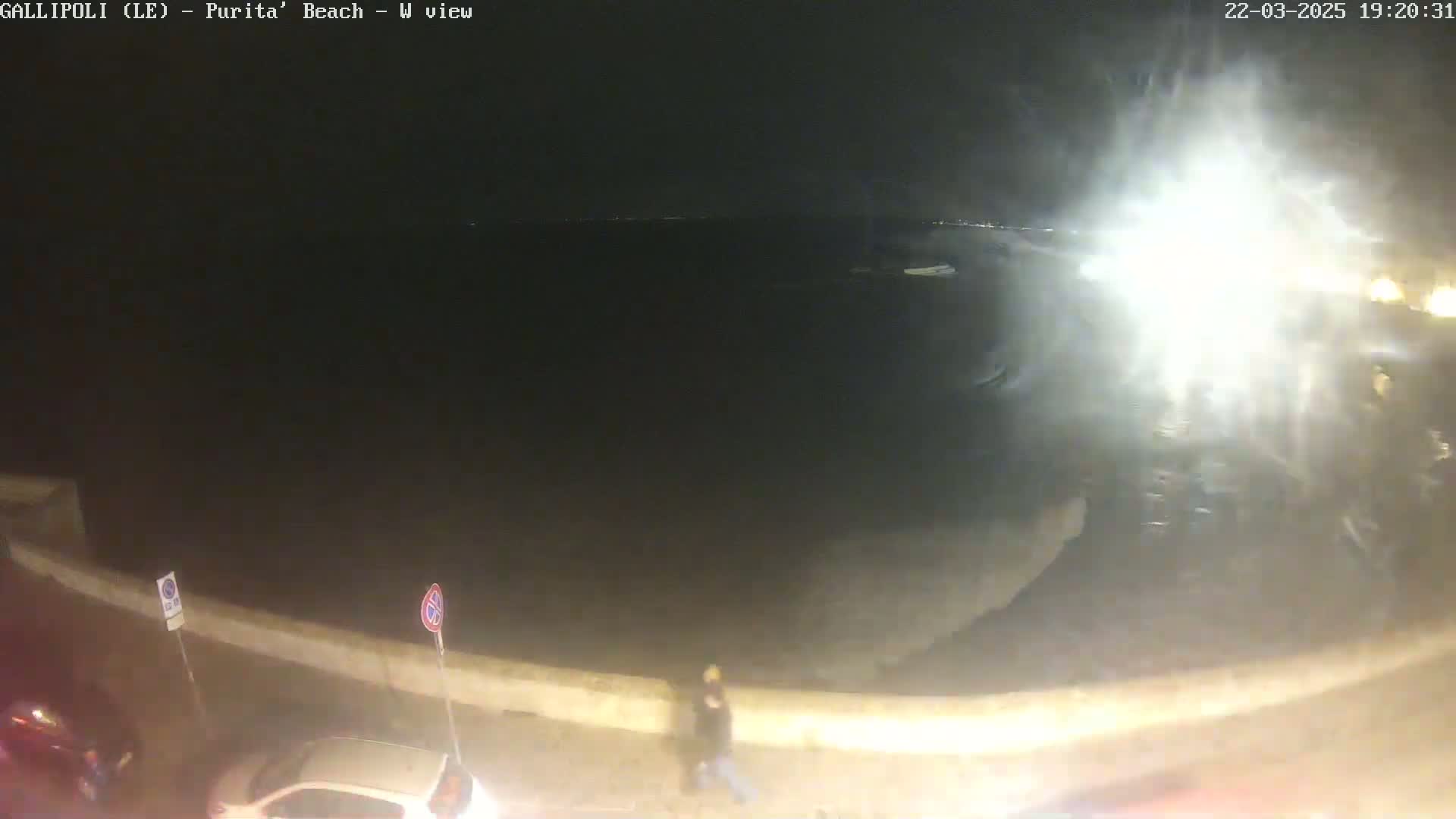 A nighttime, seaside view shows a dark, choppy sea under a cloudy sky, with a person walking along a wall next to parked cars.