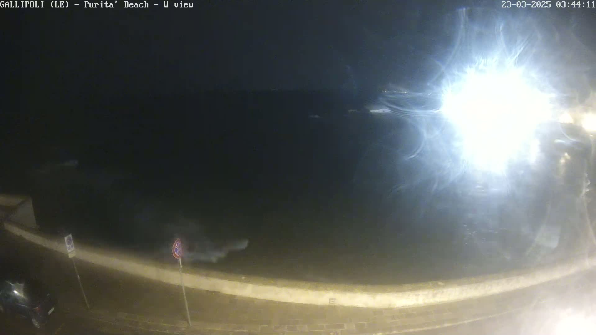 A nighttime view of a dark ocean with bright light reflecting off the water from a source out of frame, seen from a low vantage point near a seawall.