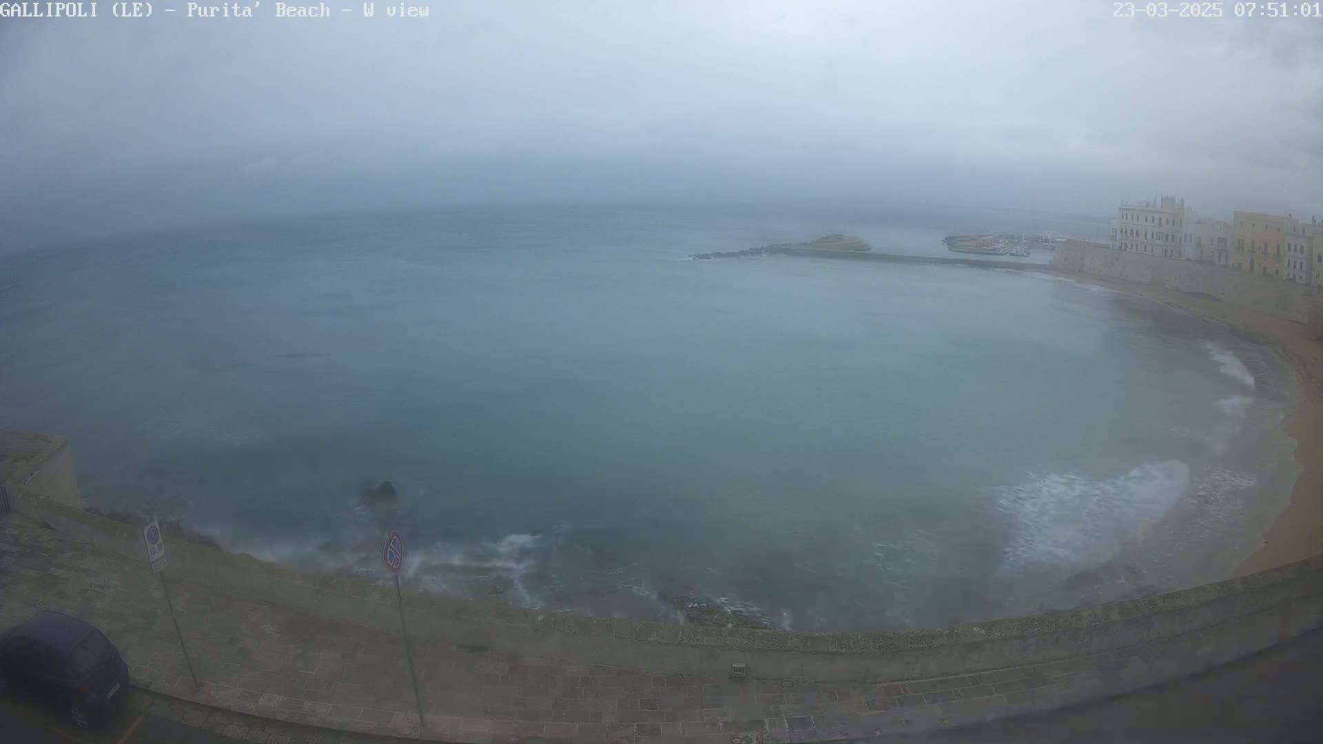 A hazy, overcast day reveals a coastal scene with a calm sea lapping against a stone embankment and buildings in the background.