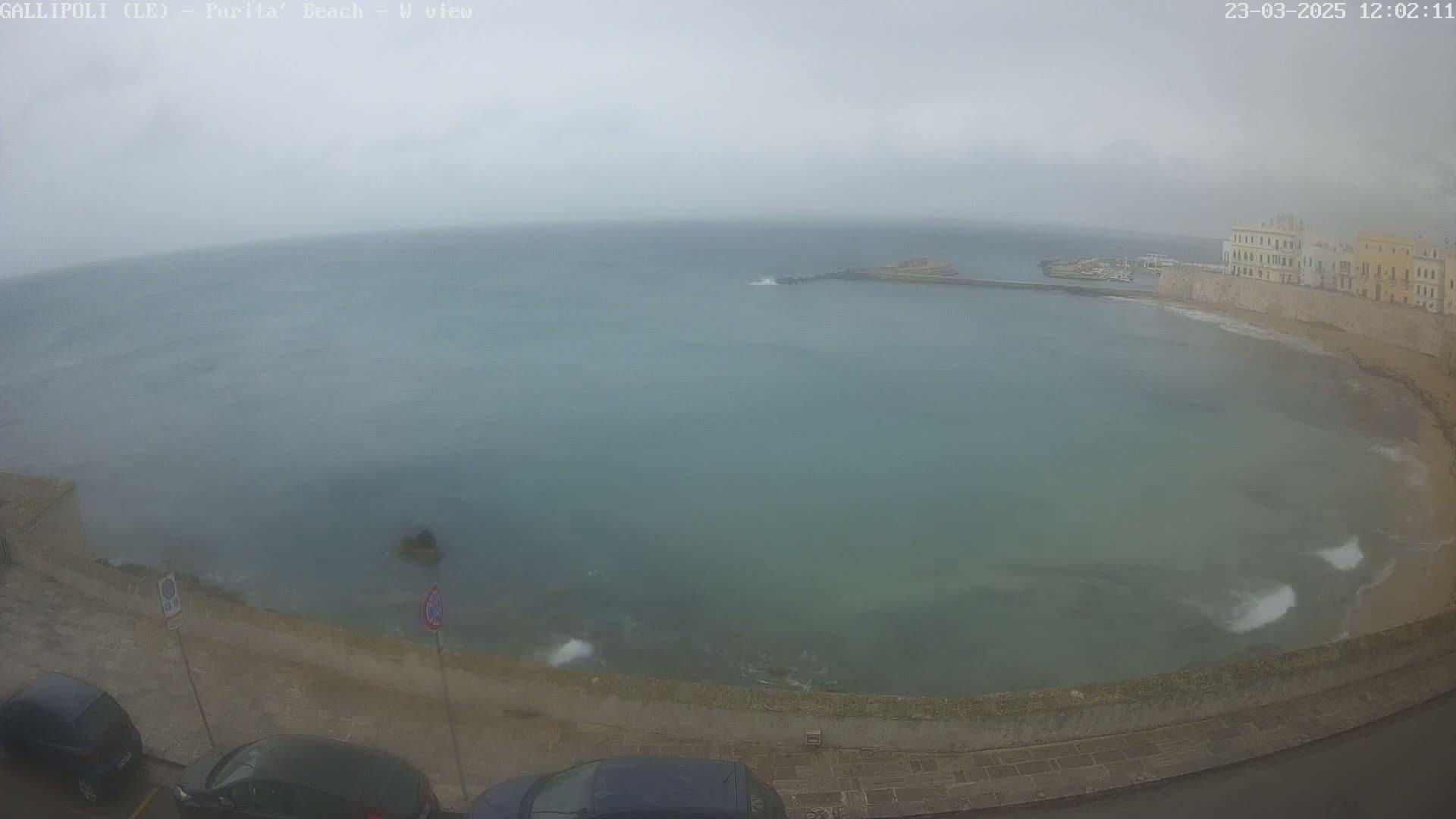 A seaside town with overcast skies and choppy water is viewed from above, showing parked cars along a paved road next to a stone wall bordering the shore.