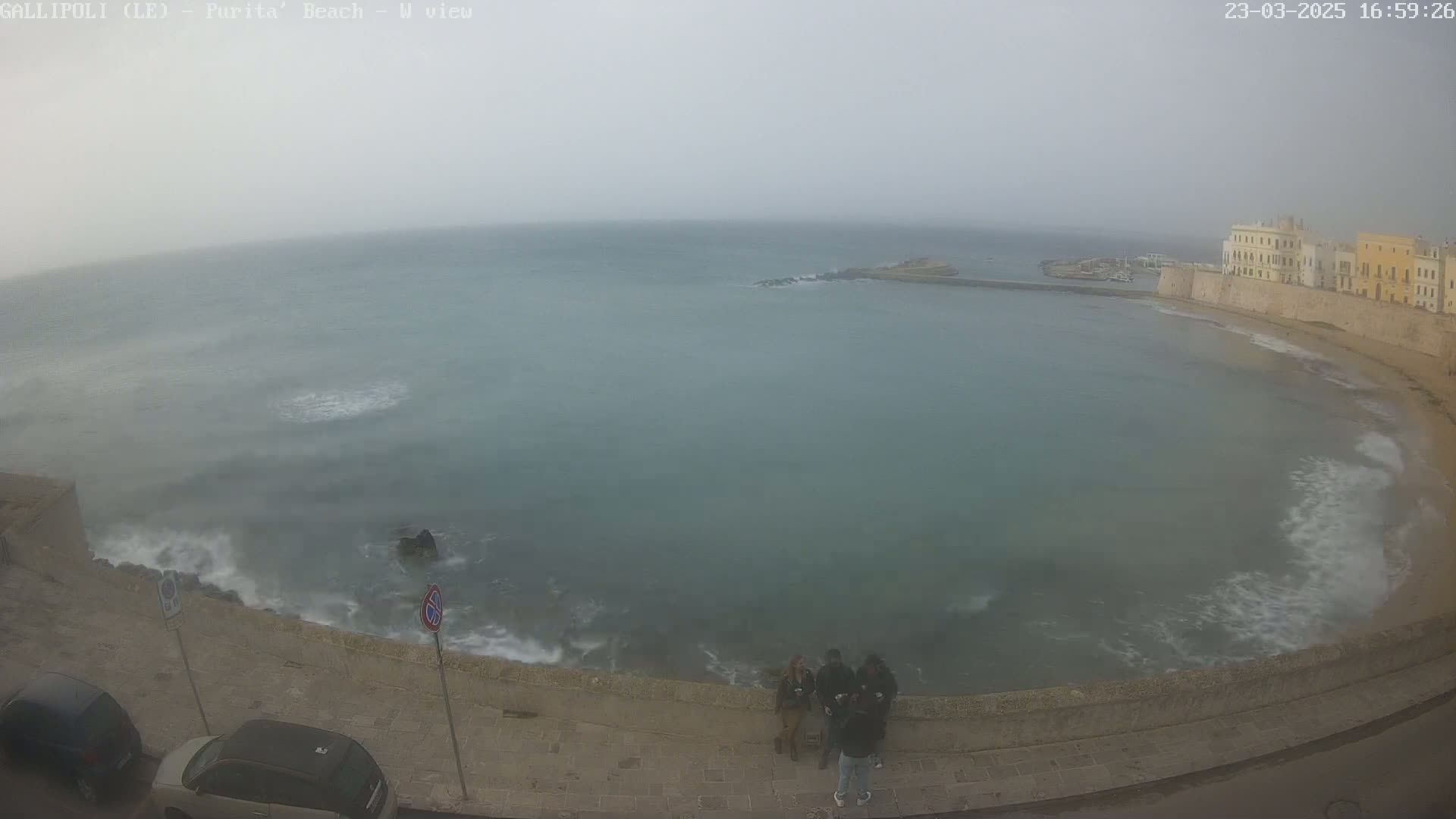 A hazy, overcast day shows a coastal scene with a calm, grayish-green sea lapping at a stone wall, where several people stand near parked cars.