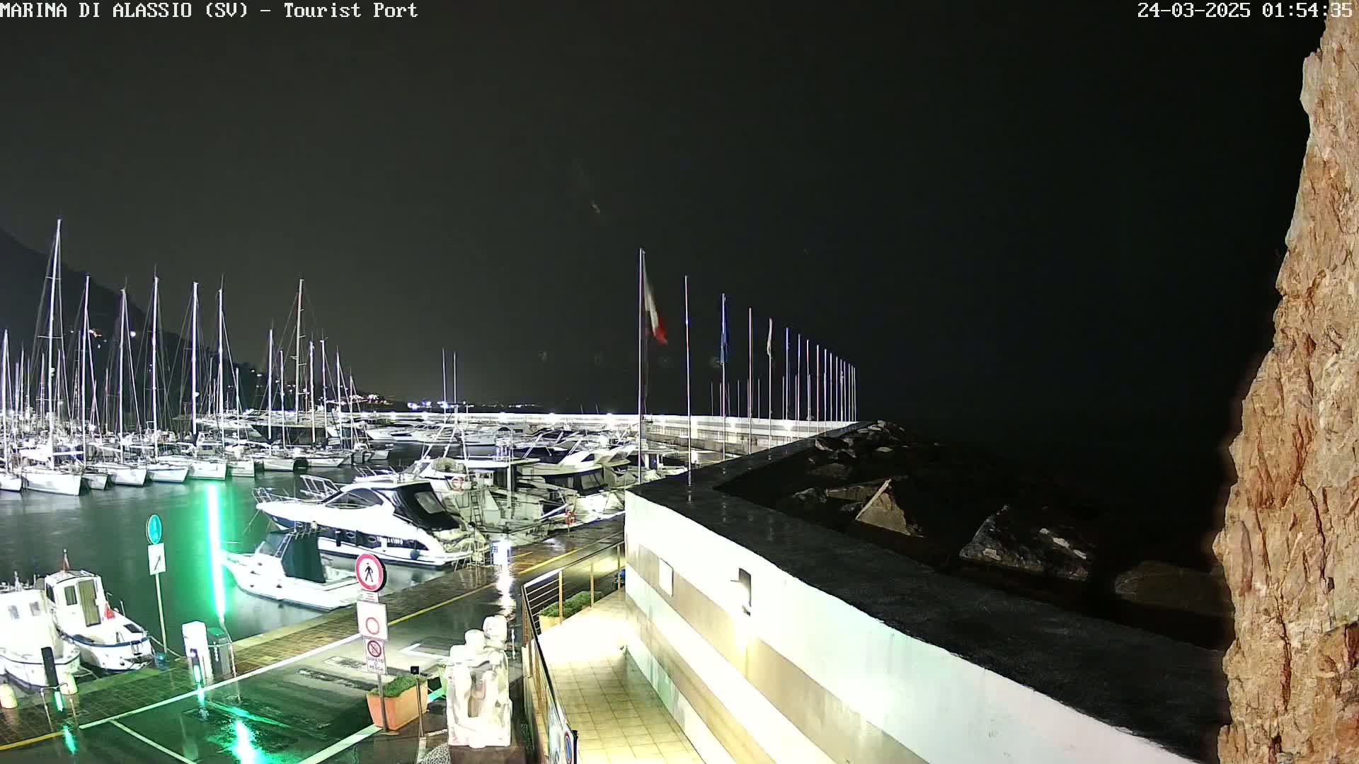 A nighttime view of a marina with numerous boats docked, under a dark sky with rain.