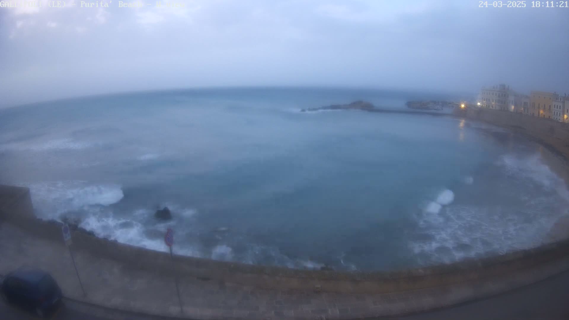 A hazy, overcast day shows a partly enclosed body of water with waves crashing against a seawall next to buildings.