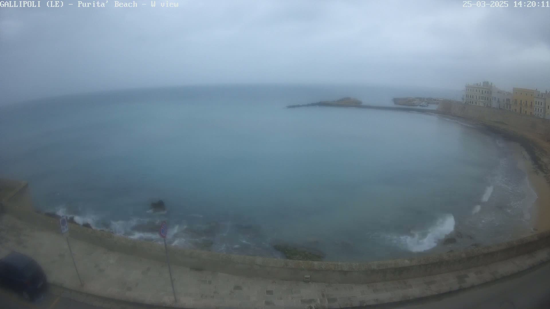 A hazy, overcast day reveals a coastal scene with a calm bay partially enclosed by a stone wall, buildings, and a small beach.